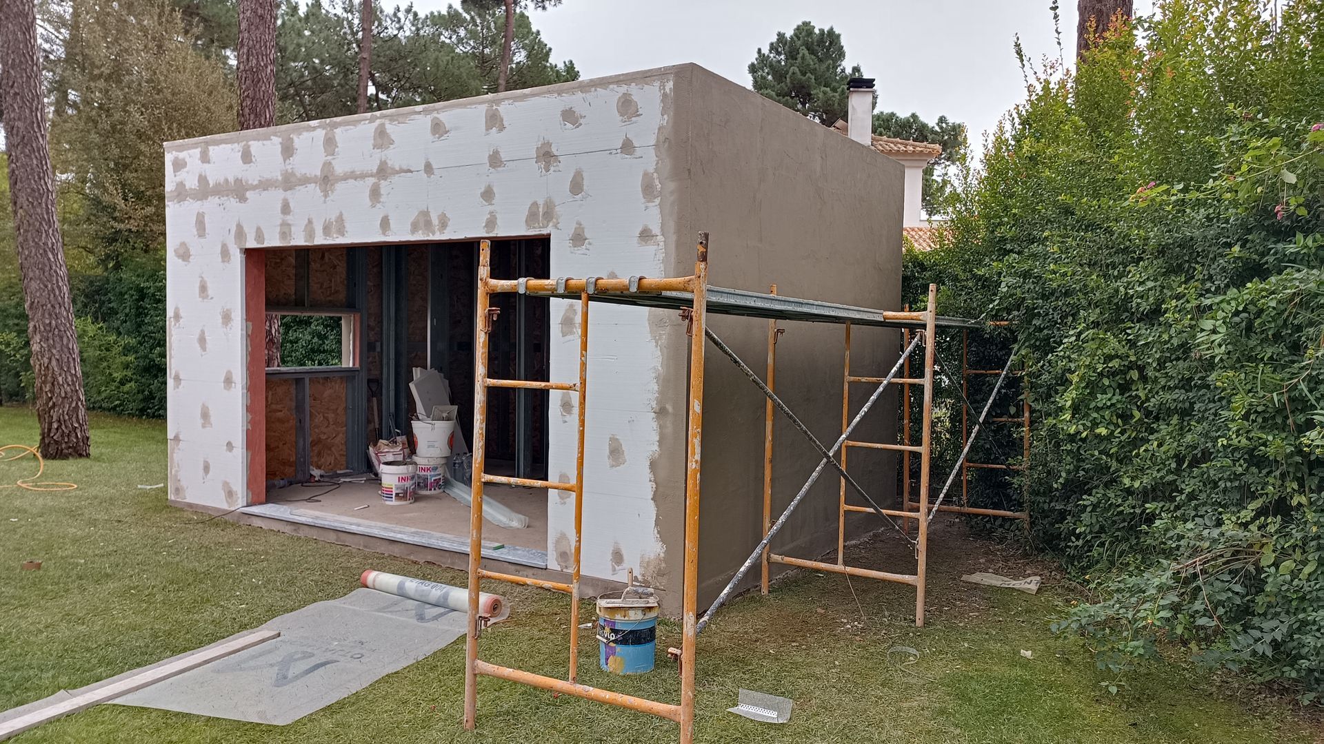 Construção de um pequeno edifício retangular; andaimes na frente, rodeado por grama verde e árvores.