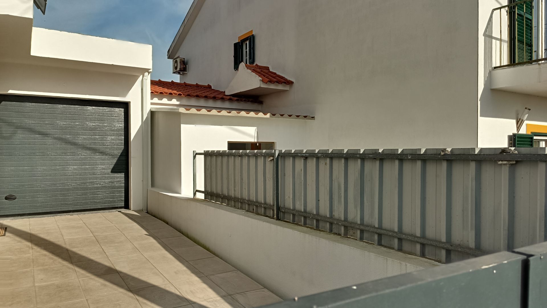Prédio branco com porta de garagem cinza, cerca de metal e telhado de telhas vermelhas sob um céu azul.