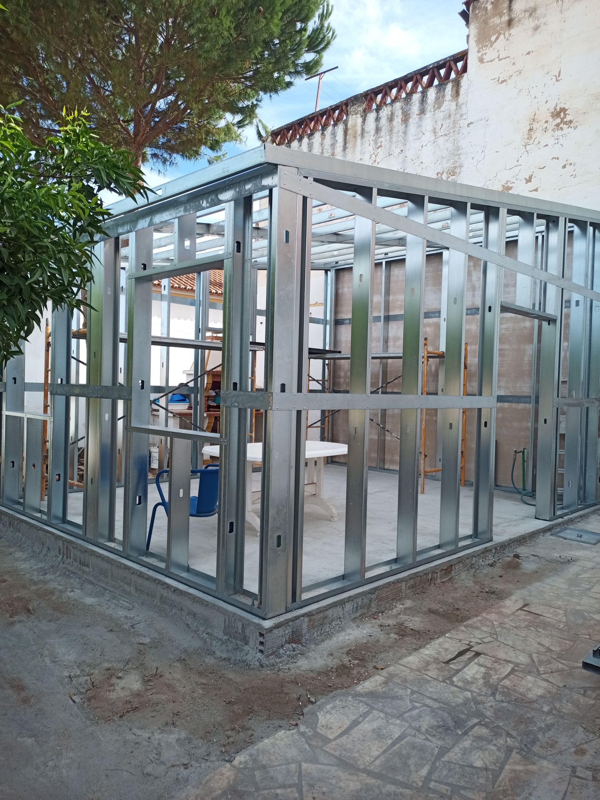 Estrutura metálica para as paredes de um edifício em construção, ao ar livre, perto de um prédio deteriorado e de uma árvore.