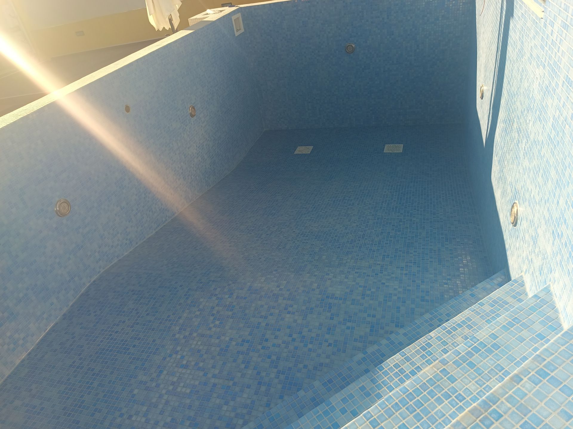 Piscina vazia revestida com azulejos azuis, com degraus e luzes, vista de cima.