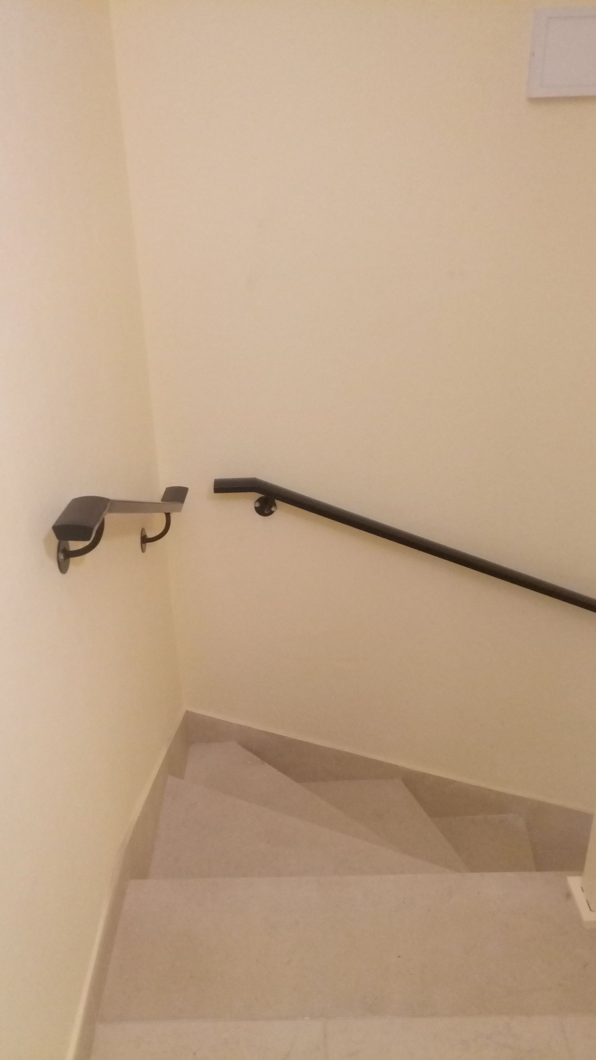 Escada com corrimão preto ao longo de uma parede cor creme, que desce para o andar inferior. O corrimão termina em um gancho na parede do canto.