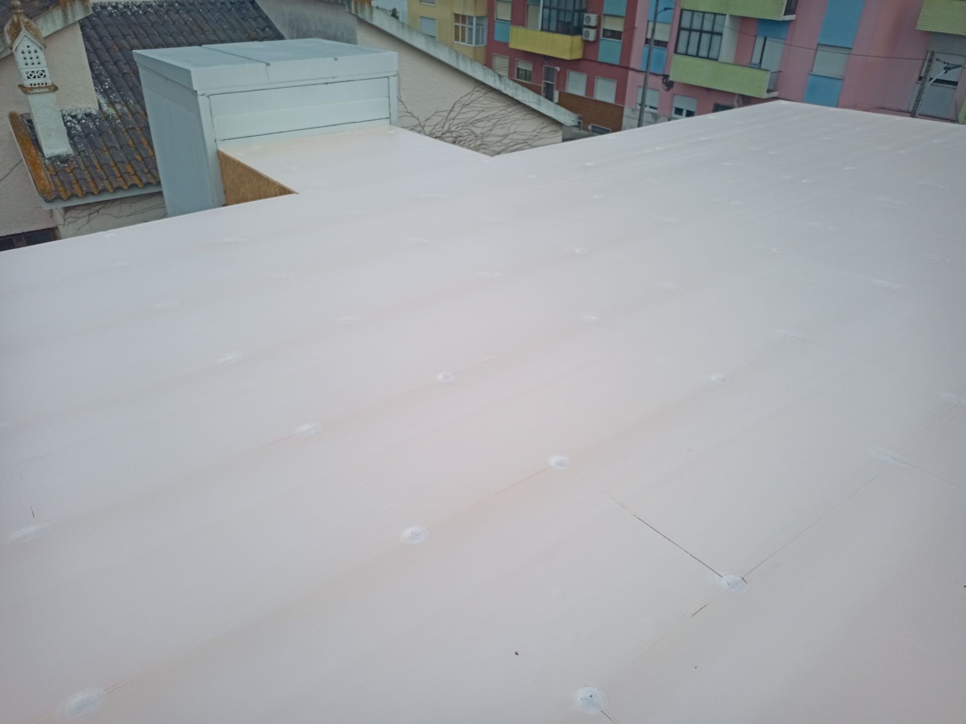 Telhado plano e claro com fixadores visíveis. Prédios e rua ao fundo.