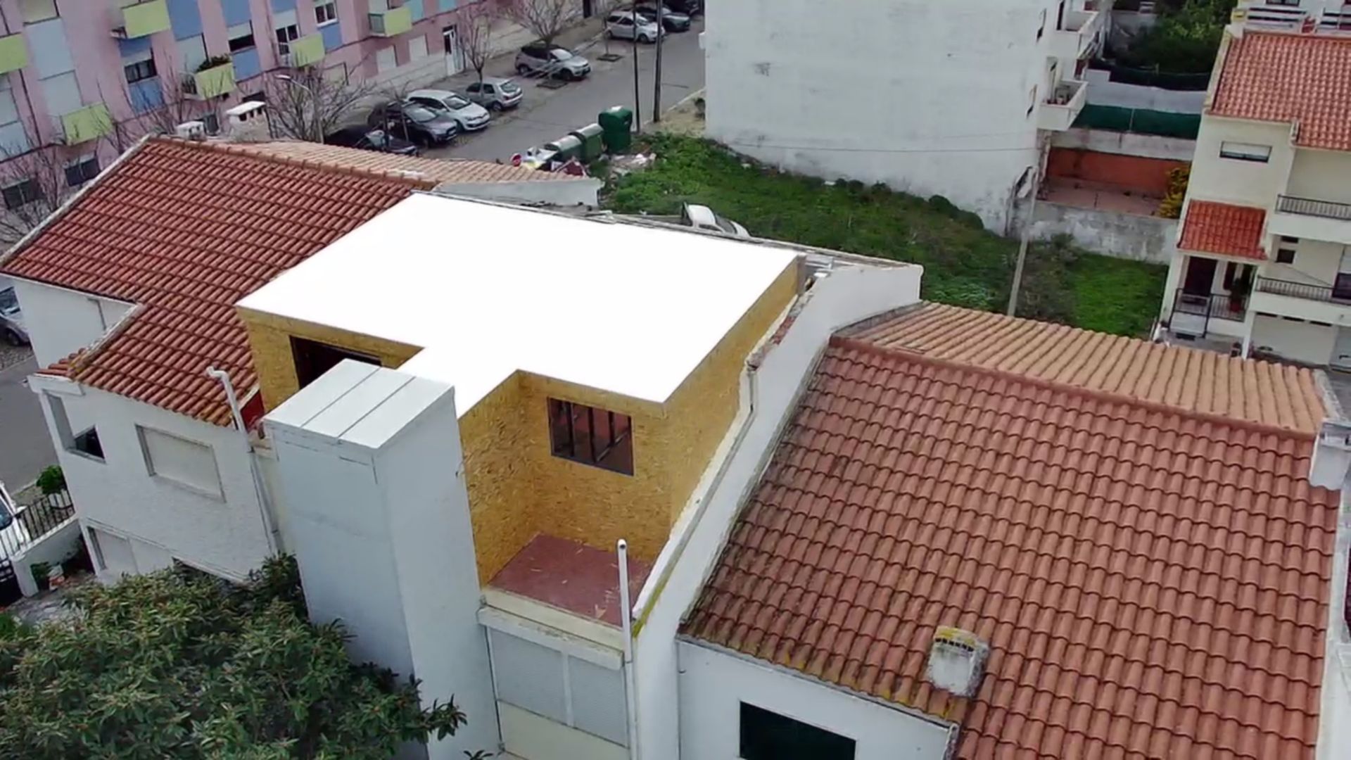 Vista do telhado de várias casas com telhados de telha laranja, um telhado plano branco e um prédio amarelo com uma varanda a céu aberto.