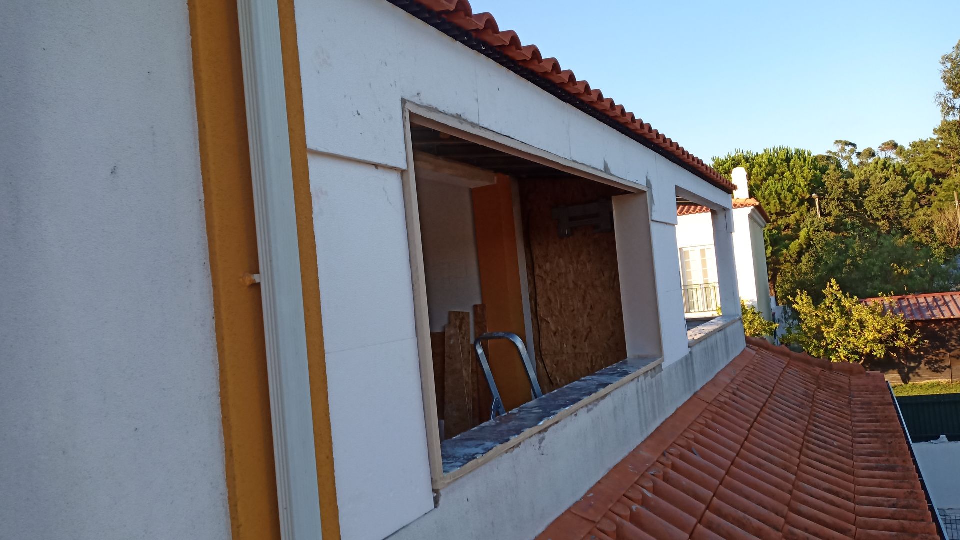 Vista exterior de um edifício com uma janela faltando, coberta com tábuas de madeira. Telhado laranja e detalhes em amarelo.