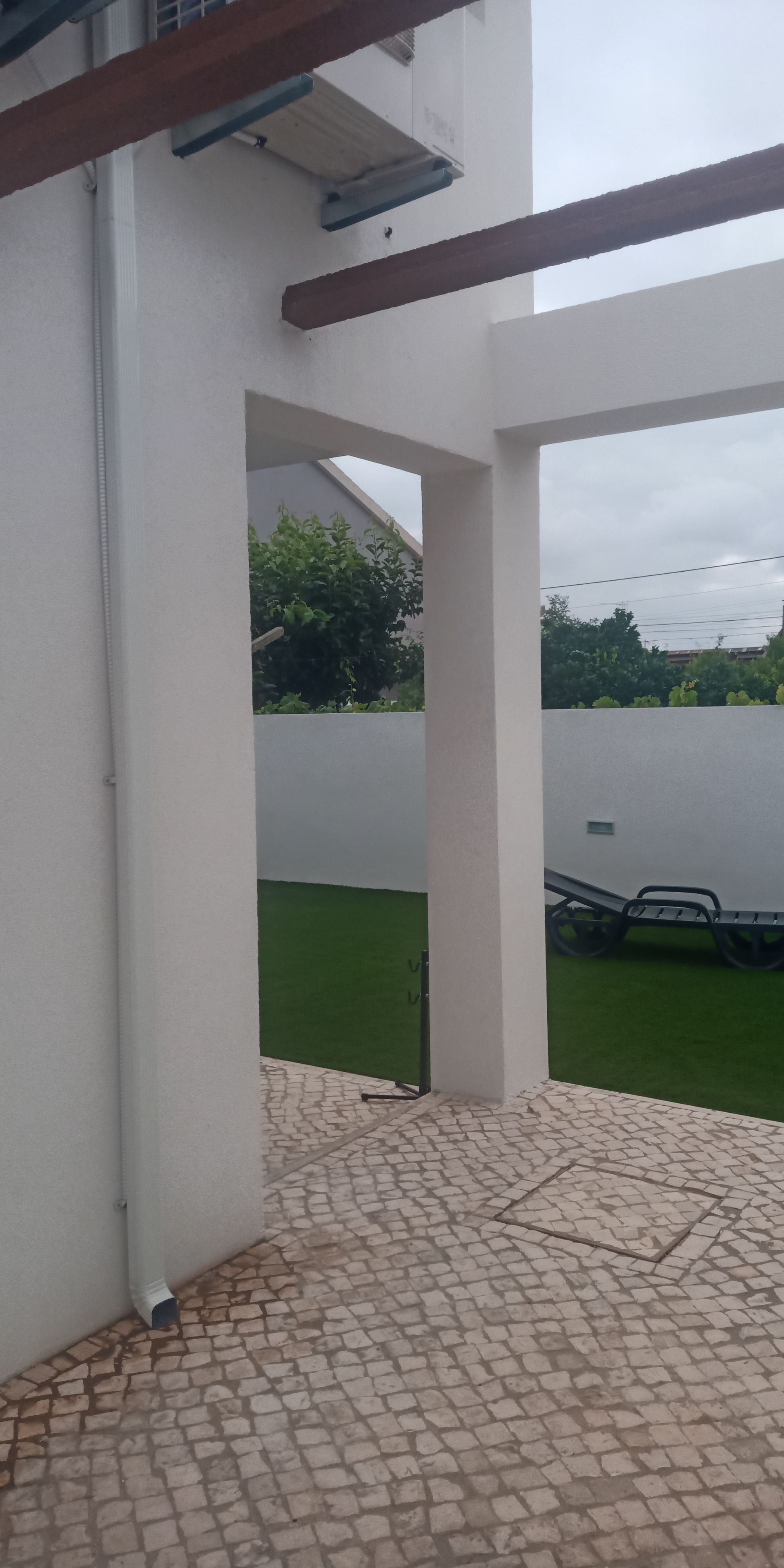 Vista exterior de um edifício com colunas de sustentação brancas, vigas de madeira e um pátio de tijolos com vista para a grama verde e uma cerca branca.