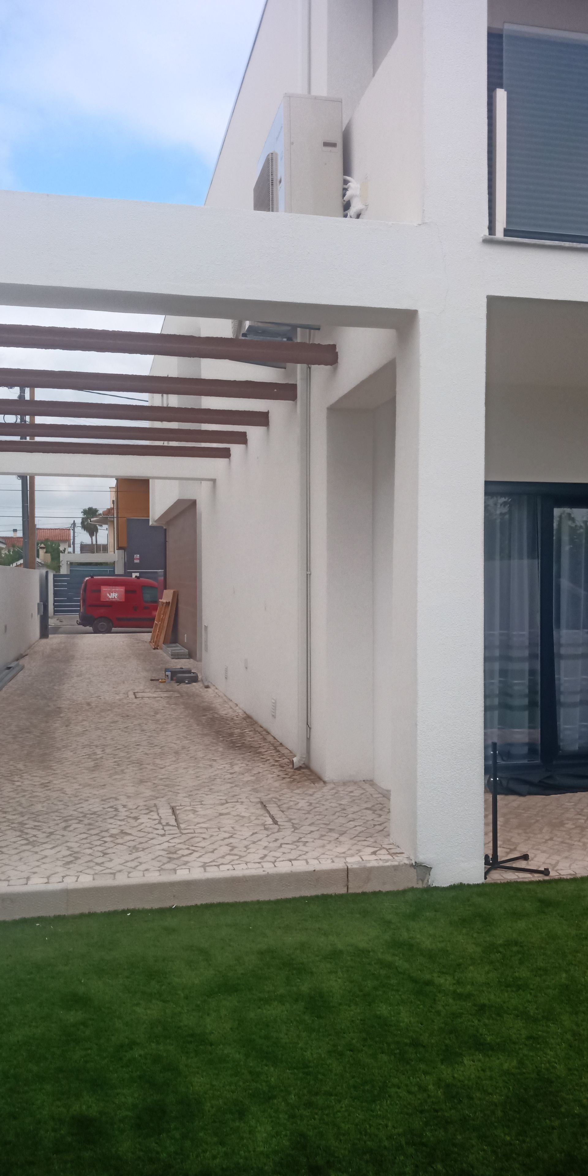 Prédio moderno branco com pérgola e entrada de automóveis em tijolo, grama verde em primeiro plano.