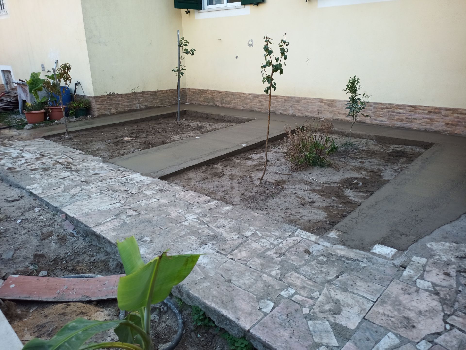 Pátio com caminho de pedra e calçadas de concreto recém-construídas, pequenas árvores e um canteiro de jardim.