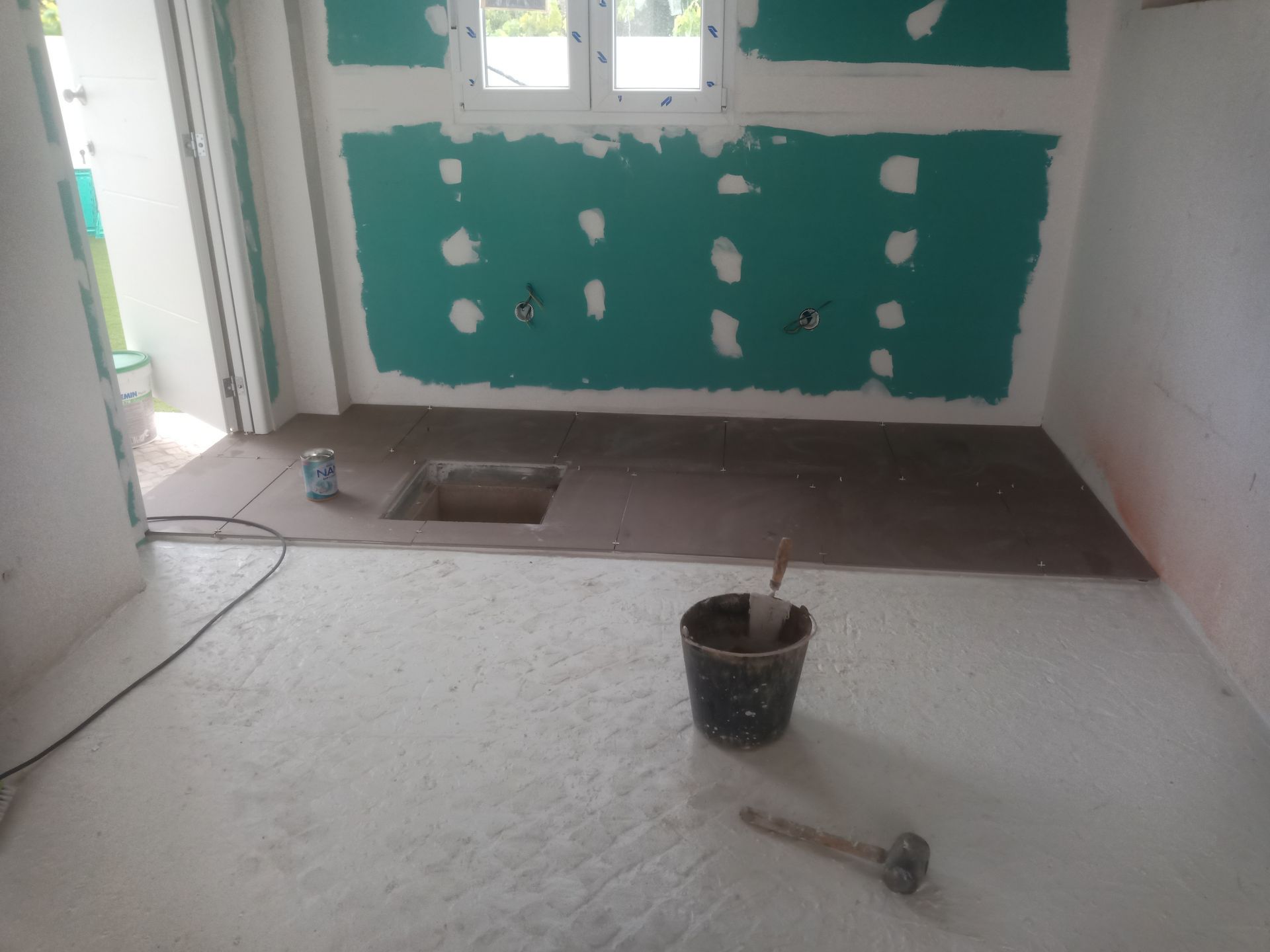 Banheiro em construção: parede verde, detalhe escuro no piso, contrapiso branco, balde, martelo.