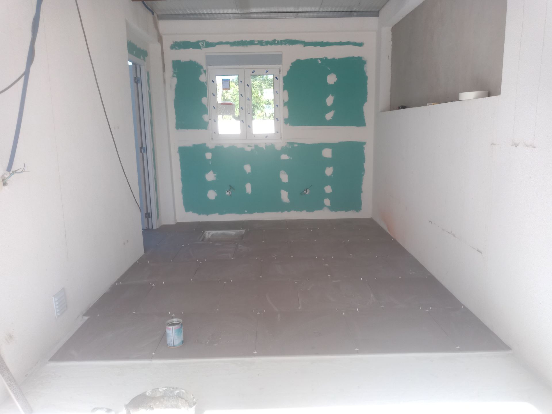 Interior de um cômodo em construção; drywall verde ao redor de uma janela, piso cinza, paredes brancas.