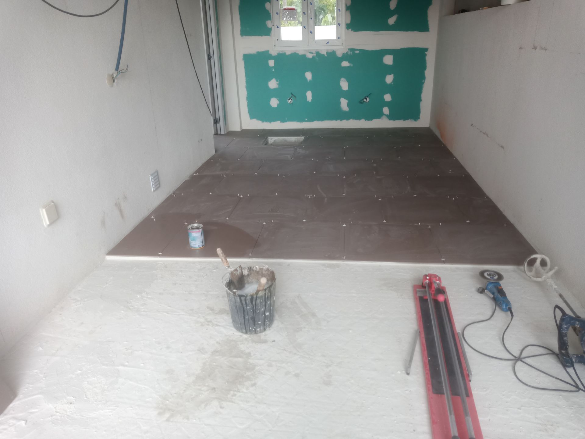 Quarto em reforma; trabalho de azulejamento em andamento. Azulejos cinza assentados, ferramentas, baldes e paredes parcialmente pintadas.