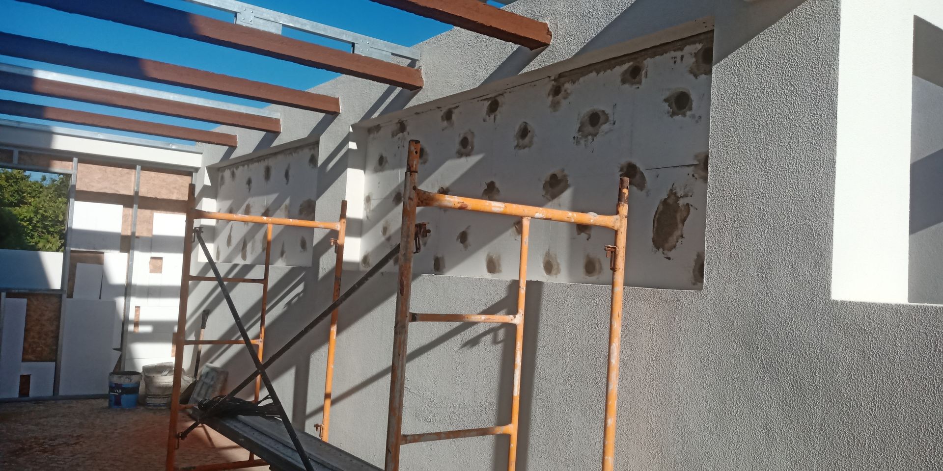 Canteiro de obras com andaimes e isolamento de parede exposto. Vigas marrons no teto. Dia ensolarado.