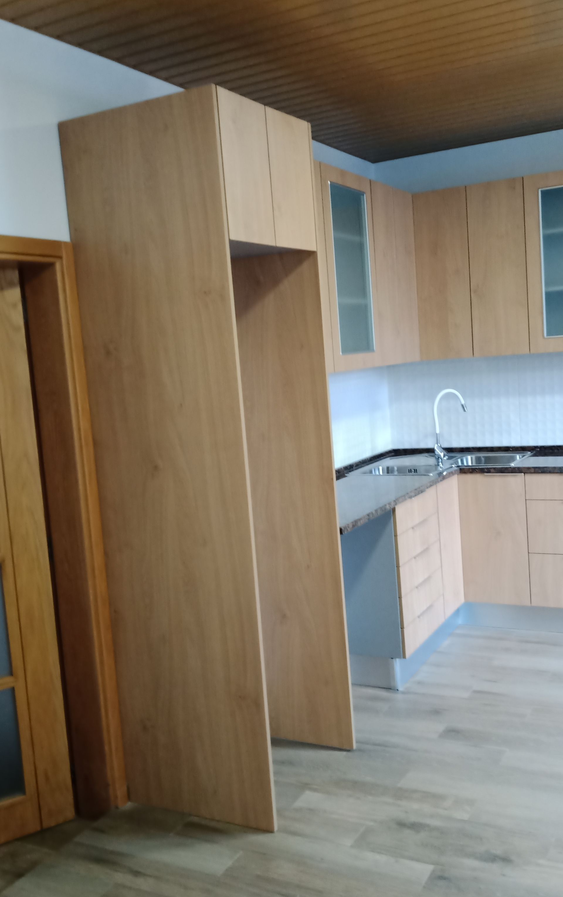 Armários de cozinha de madeira ao redor do espaço da geladeira; pia, bancada e revestimento de azulejos visíveis.