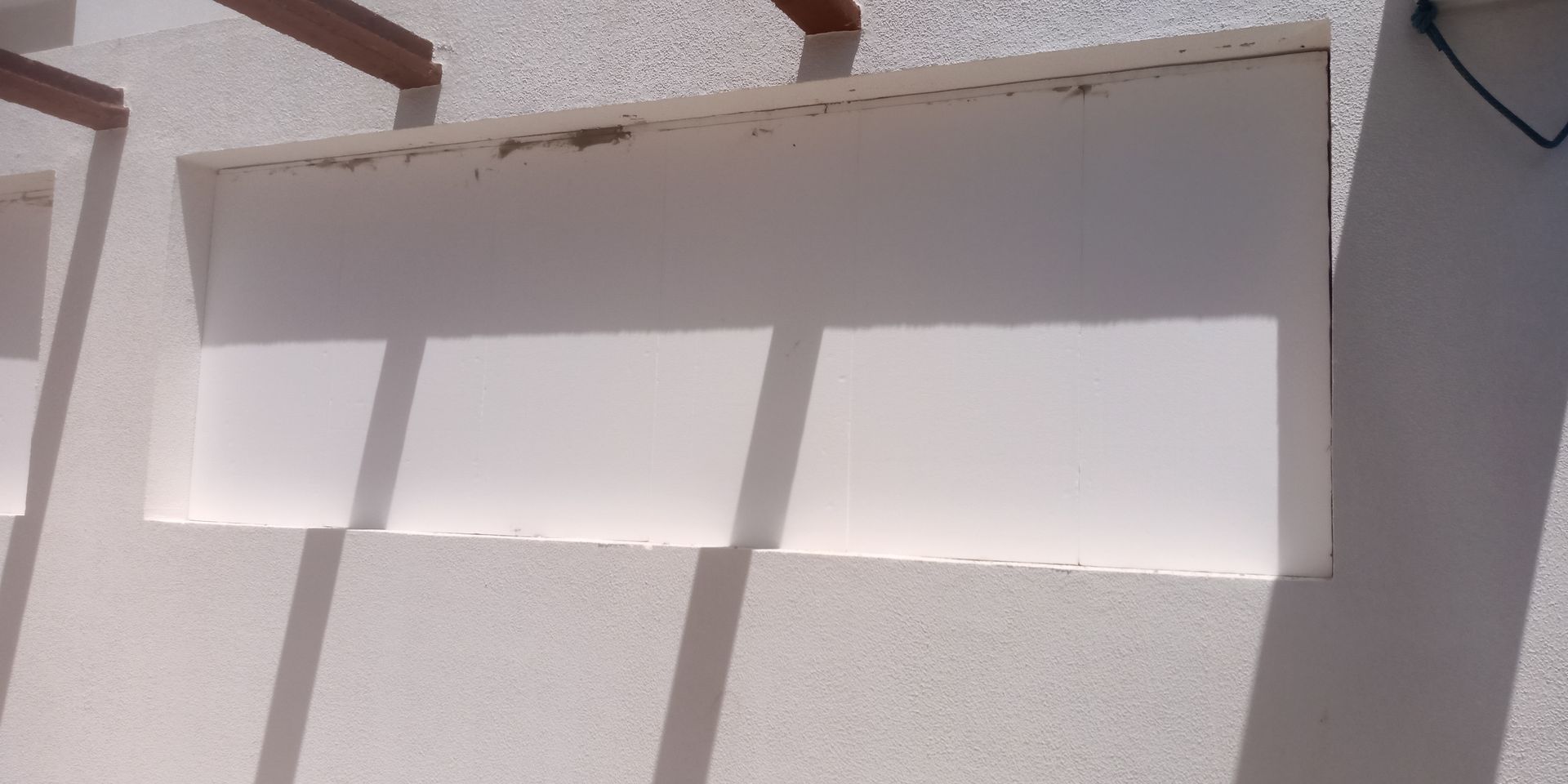 Parede branca com recortes retangulares que projetam sombras das vigas de madeira acima.