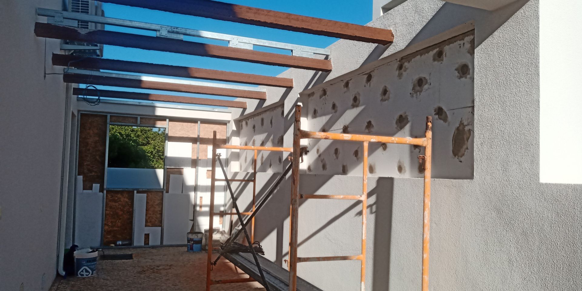 Canteiro de obras com pérgola parcialmente construída e andaimes encostados em uma parede texturizada, banhado pela luz do sol.