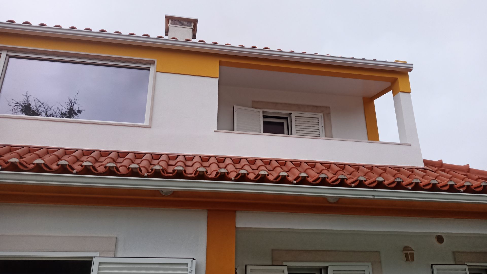 Casa de dois andares com paredes brancas, detalhes em laranja e telhado de telha vermelha. Céu nublado ao fundo.