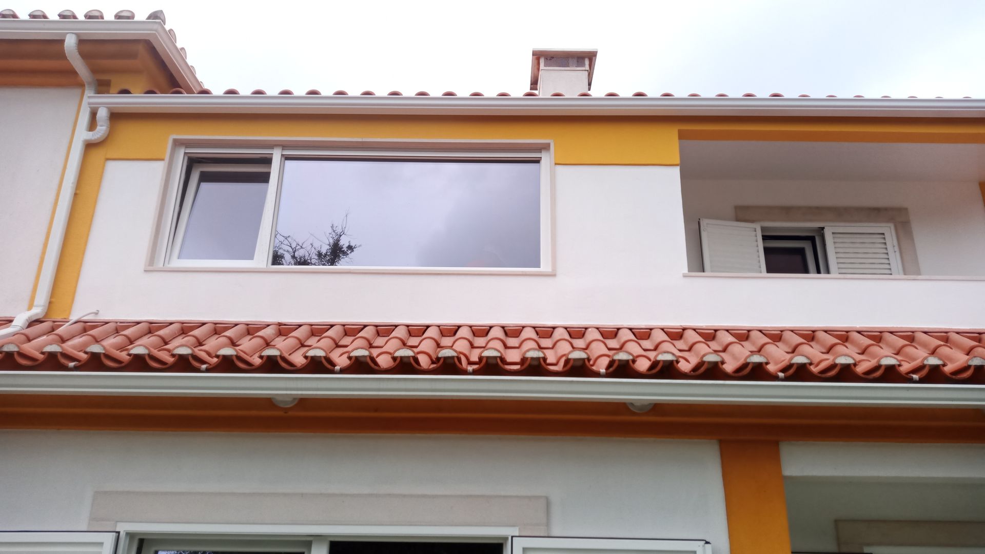 Casa de dois andares com telhado de telha vermelha, exterior branco e amarelo, janelas, varanda e calhas.