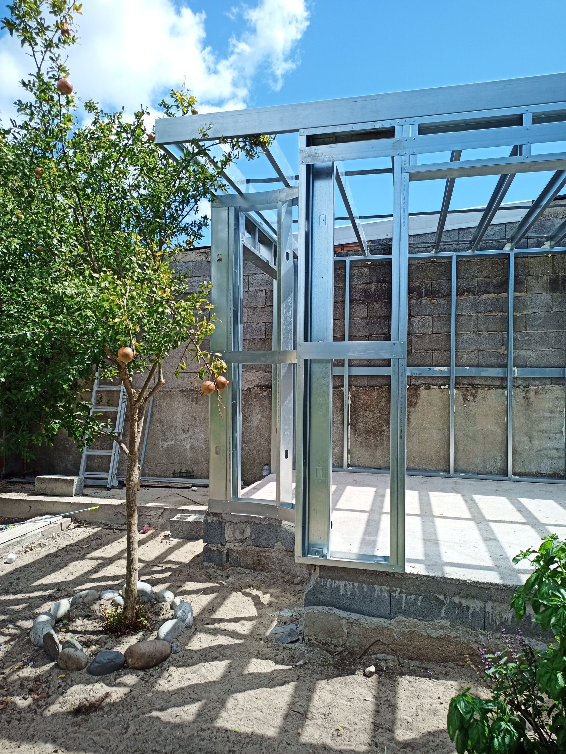 Estrutura metálica de uma construção em andamento, ao lado de uma árvore e um muro de pedra, ao ar livre em um dia ensolarado.