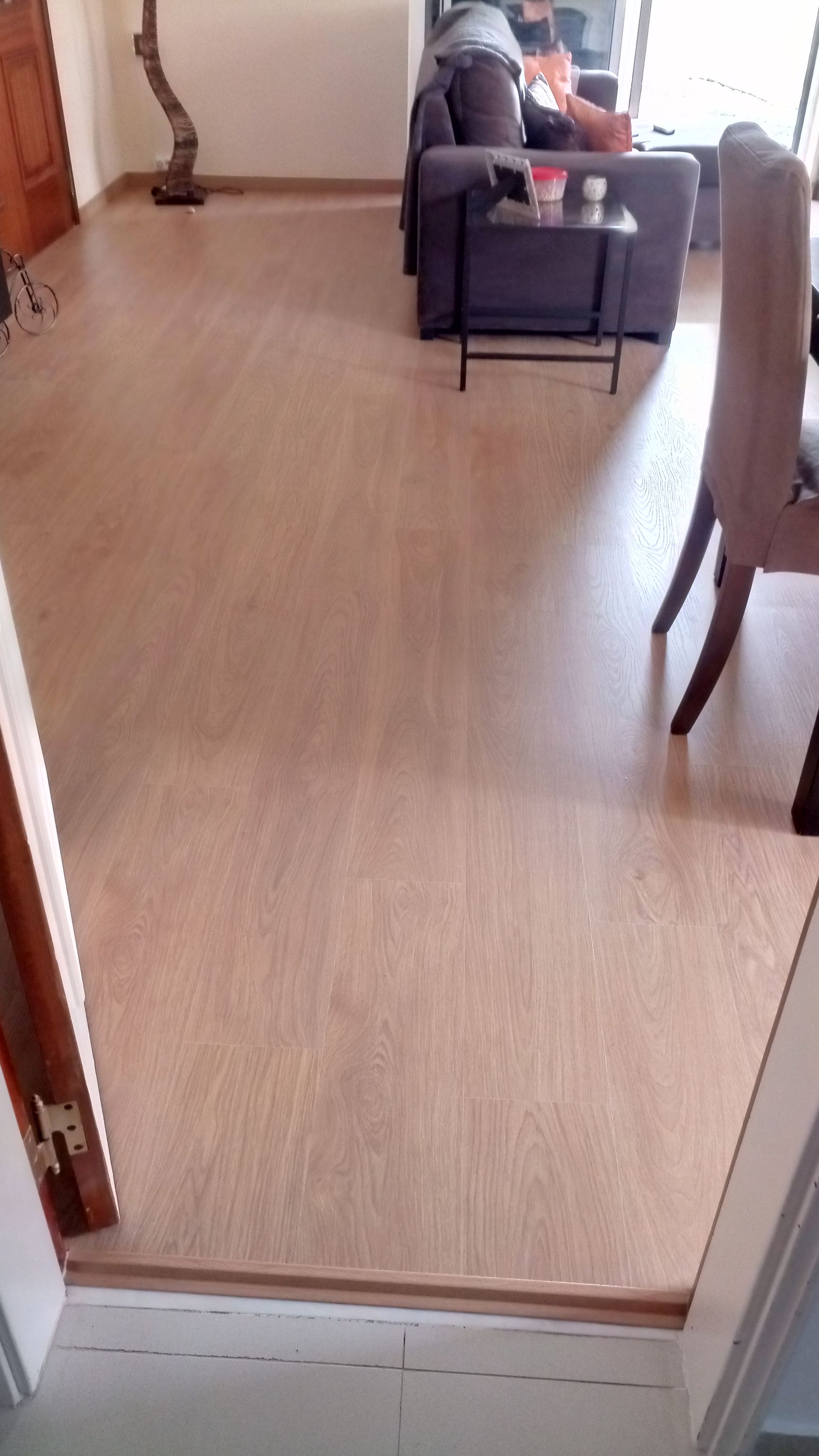 Piso de madeira maciça marrom-claro, que leva da porta de entrada até uma sala de estar com sofá, mesa e cadeira.