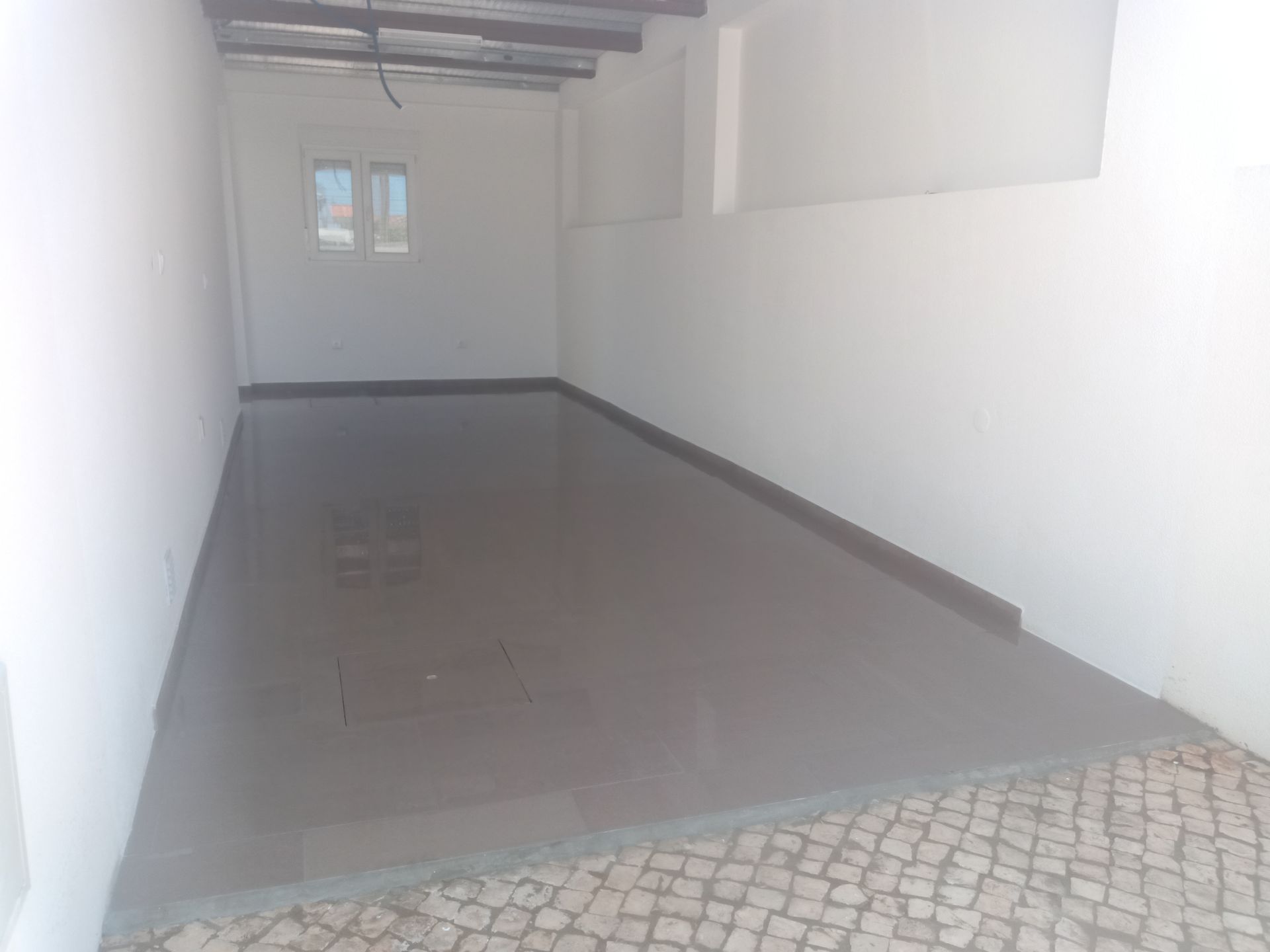 Vista interior: quarto com piso cinza recém-instalado, paredes brancas, janela pequena e entrada de paralelepípedos.