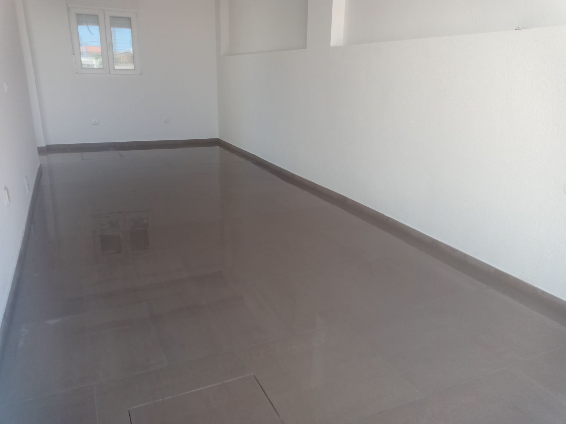 Quarto vazio com piso de azulejo cinza brilhante e paredes brancas. Pequena janela na parede do fundo.