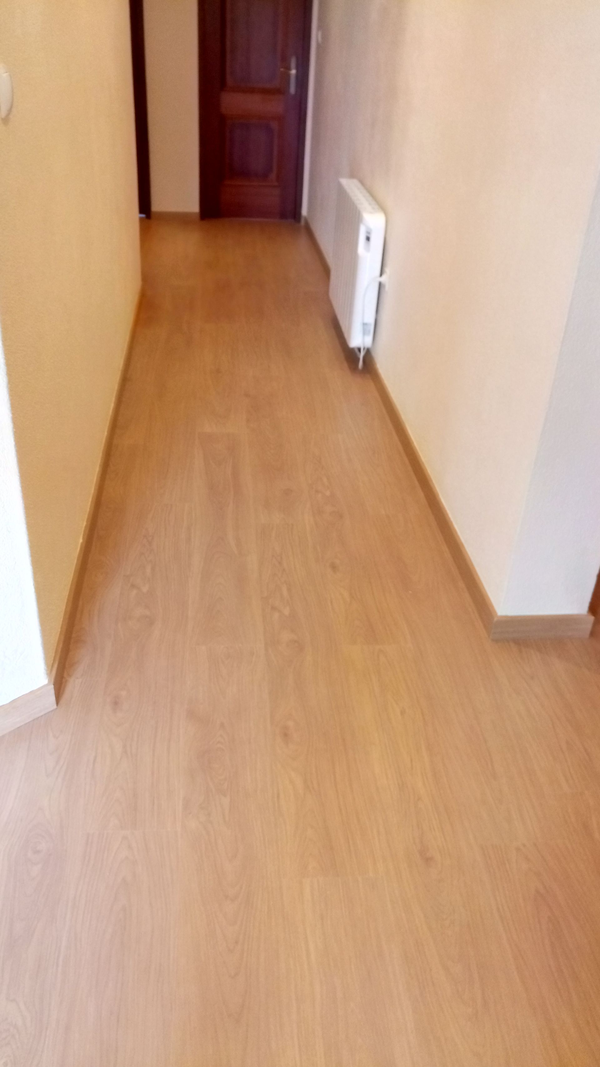 Corredor estreito com piso de madeira clara, paredes bege e um radiador branco. Uma porta marrom fechada está no final.
