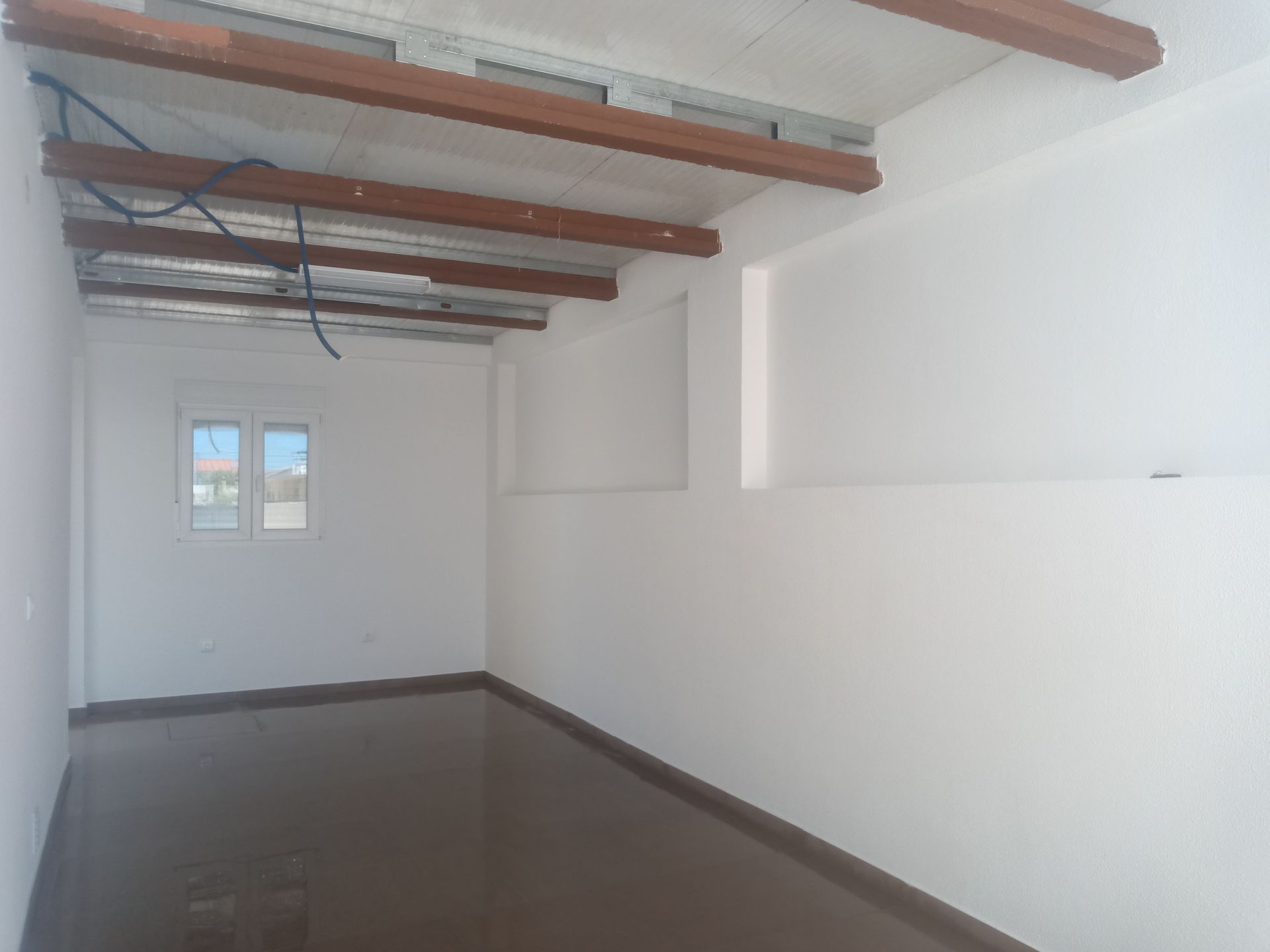 Sala vazia com paredes brancas, janela e piso escuro. Vigas de madeira sustentam o teto.