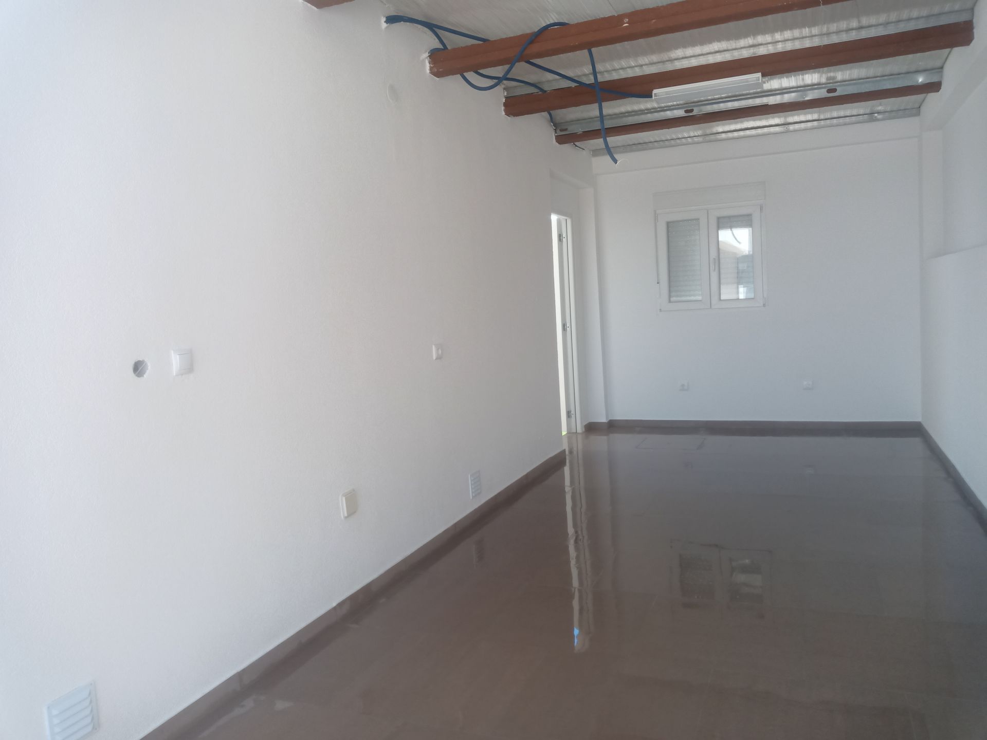 Quarto vazio com piso marrom brilhante, paredes brancas, janela pequena e vigas expostas no teto.