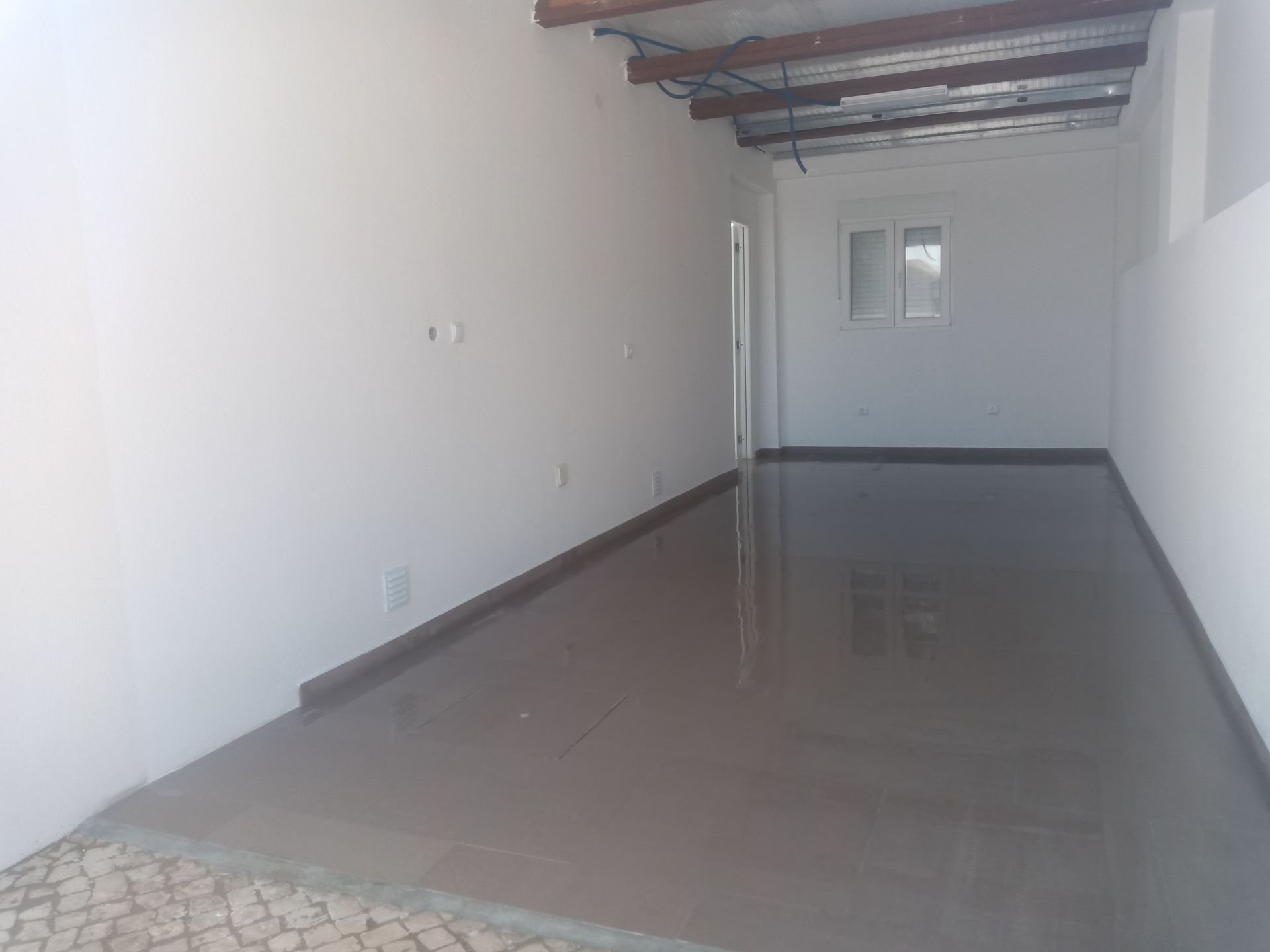 Quarto vazio com piso de cimento recém-colocado, paredes brancas, janela pequena e porta aberta.