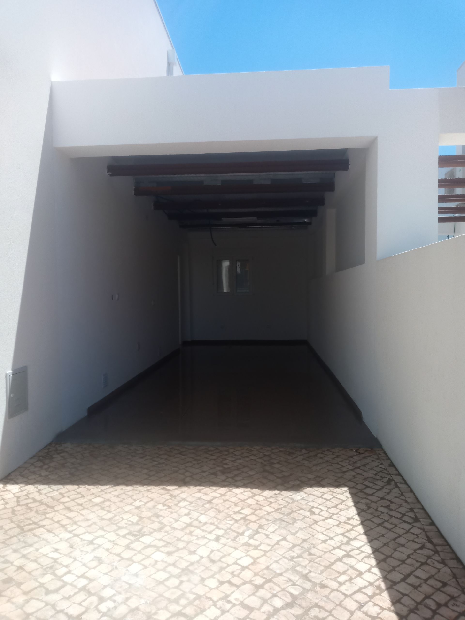 Entrada coberta para veículos com vigas escuras, paredes brancas e pavimentação em tijolo.