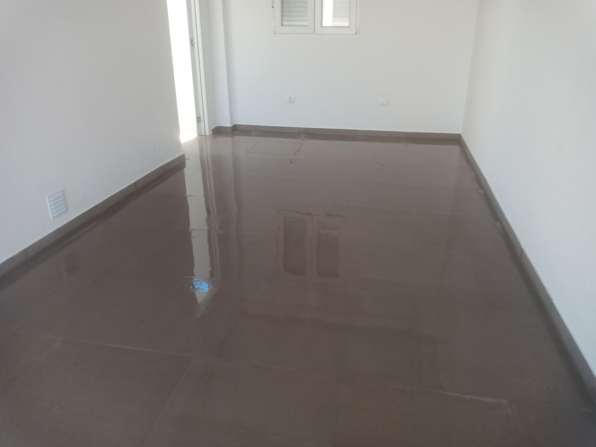 Quarto vazio com piso marrom brilhante, paredes brancas e uma pequena janela.