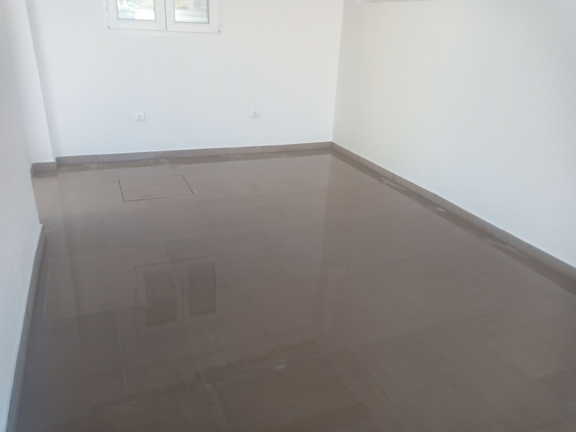 Quarto vazio com piso marrom brilhante, paredes brancas e uma pequena janela.