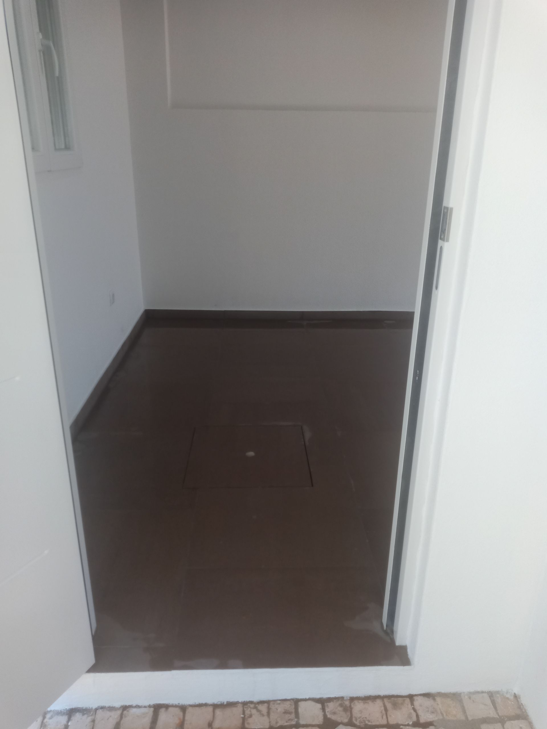 A porta se abre para um pequeno quarto com piso marrom e paredes brancas.