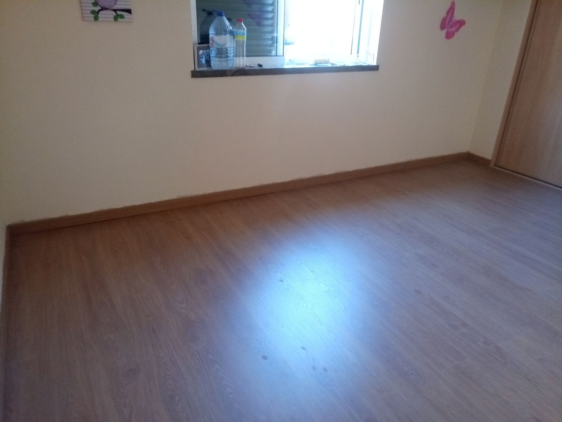 Quarto vazio com piso que imita madeira clara, paredes bege, janela com objetos no parapeito e uma porta.