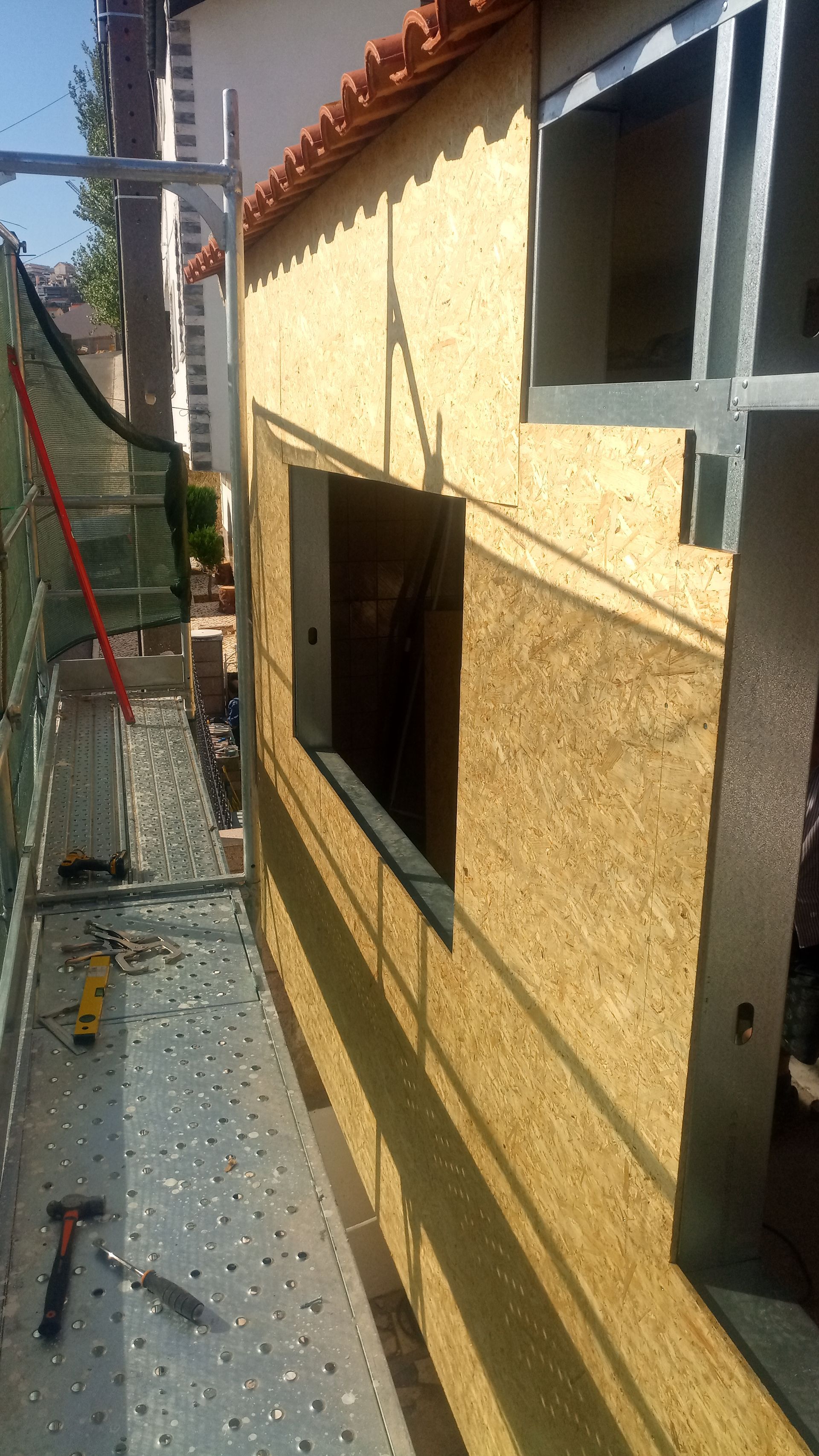Parede externa em construção, com painéis de madeira aparentes, andaimes e caixilhos de janelas.