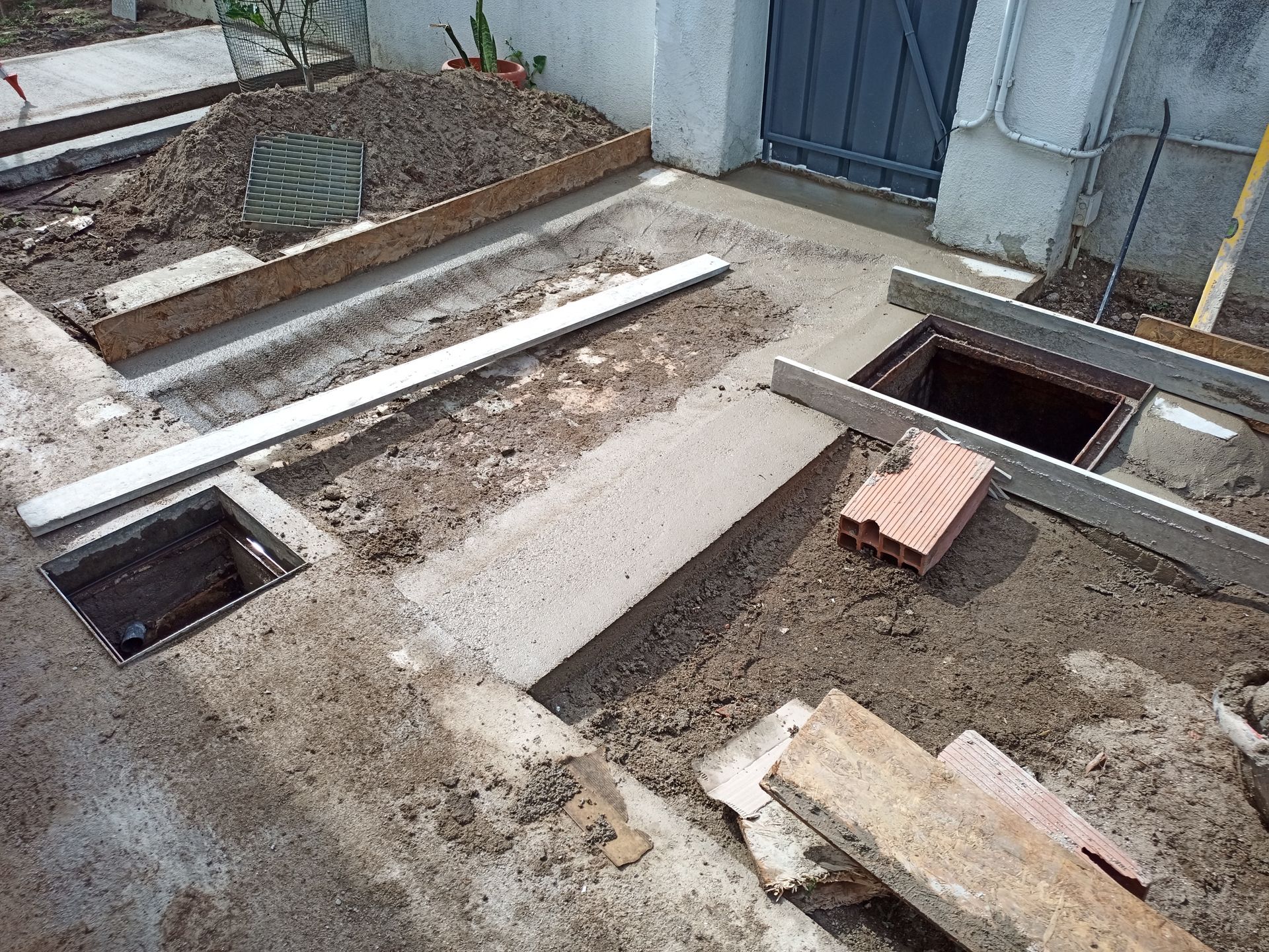 Canteiro de obras: Concreto sendo despejado ao redor de pontos de acesso de serviços públicos abertos e canteiros de terra, com ferramentas e materiais presentes.
