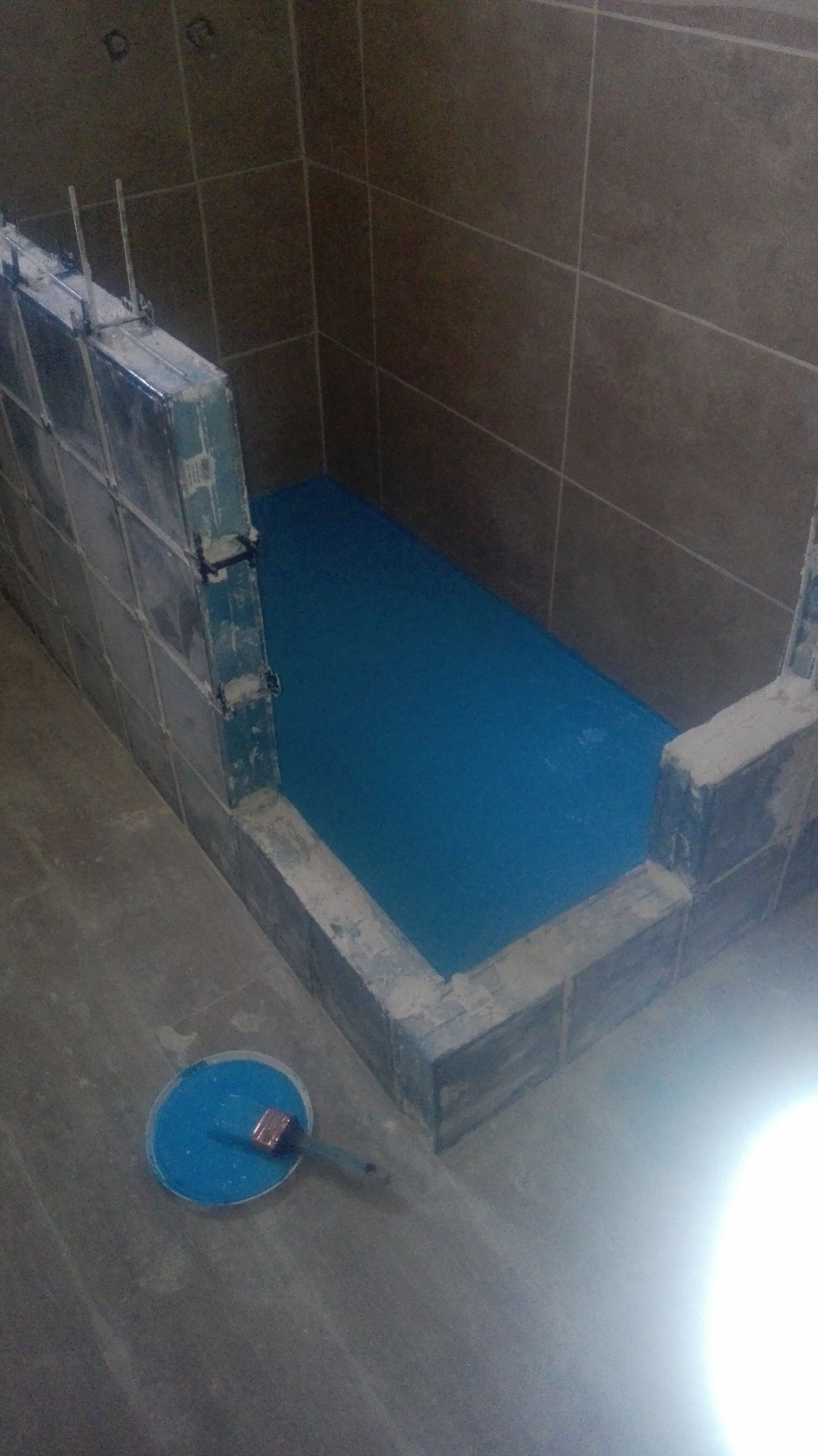 Estrutura retangular pintada de azul, possivelmente uma base de chuveiro, em construção, com paredes e um rolo de pintura por perto.