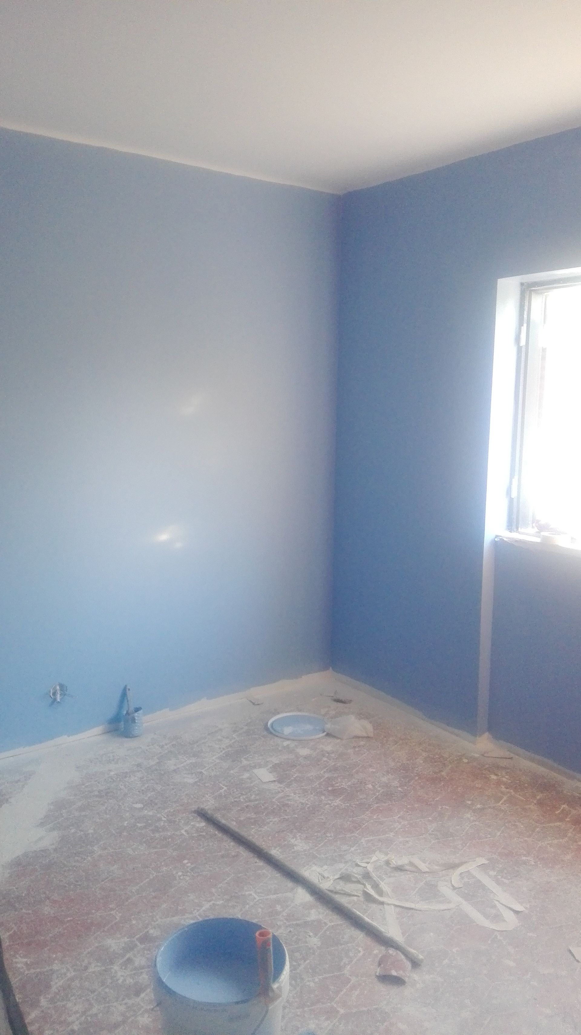 Interior de quarto pintado de azul com piso inacabado, balde e janela.