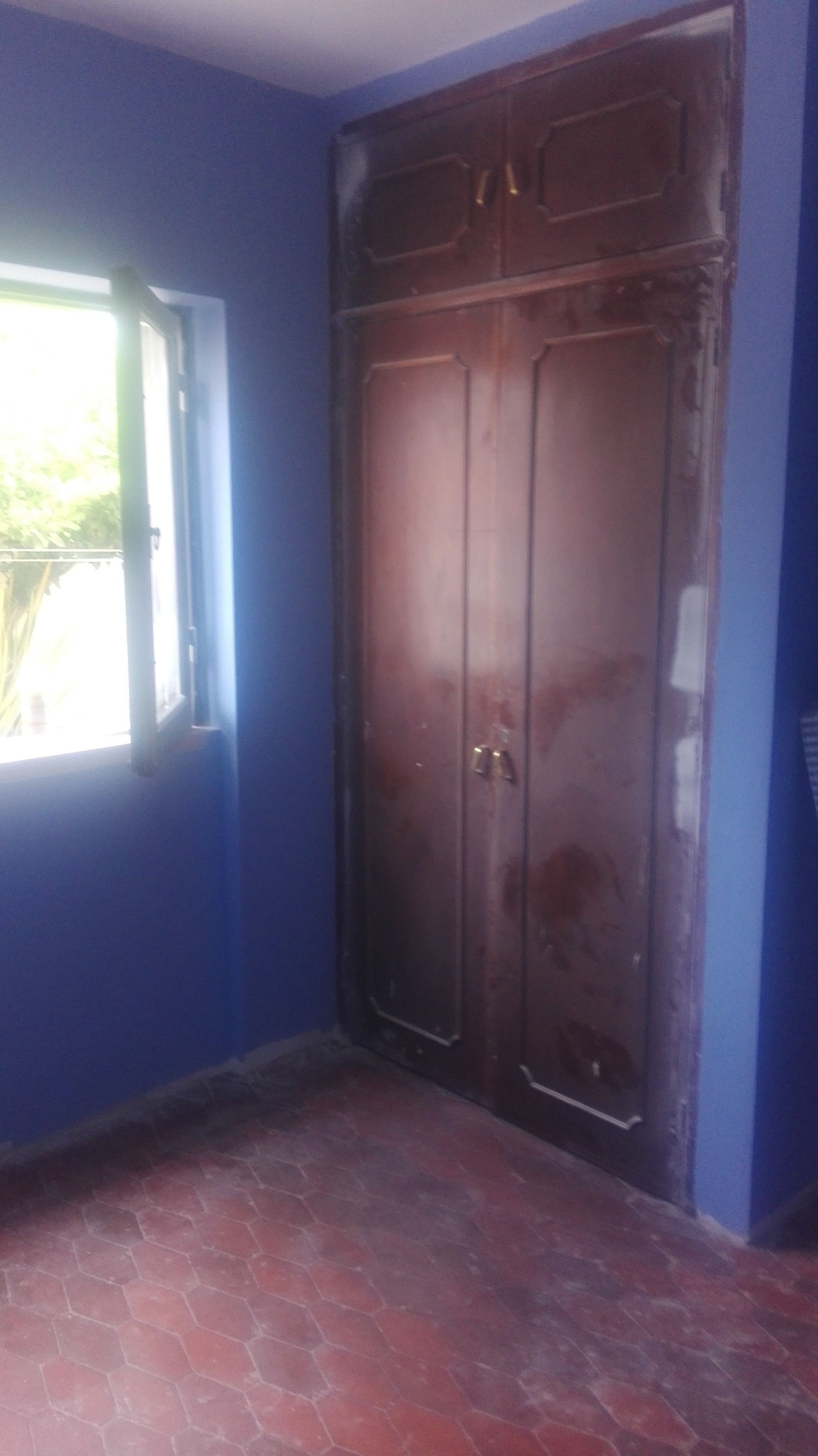 Guarda-roupa de madeira escura encostado em uma parede azul, em um quarto com janela e piso vermelho.