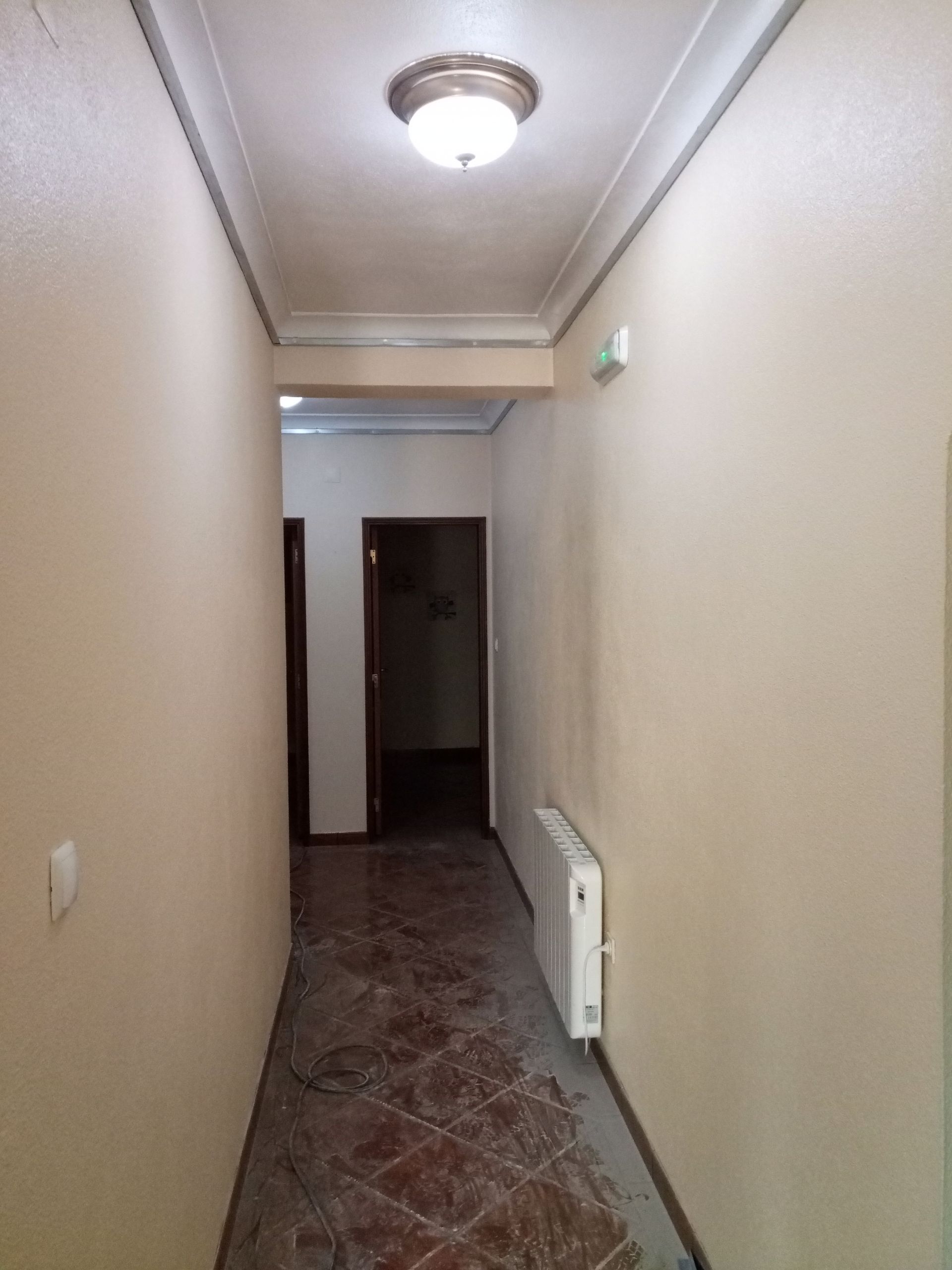 Corredor estreito com paredes bege, portas marrons e uma luminária de teto. Um radiador está à direita.