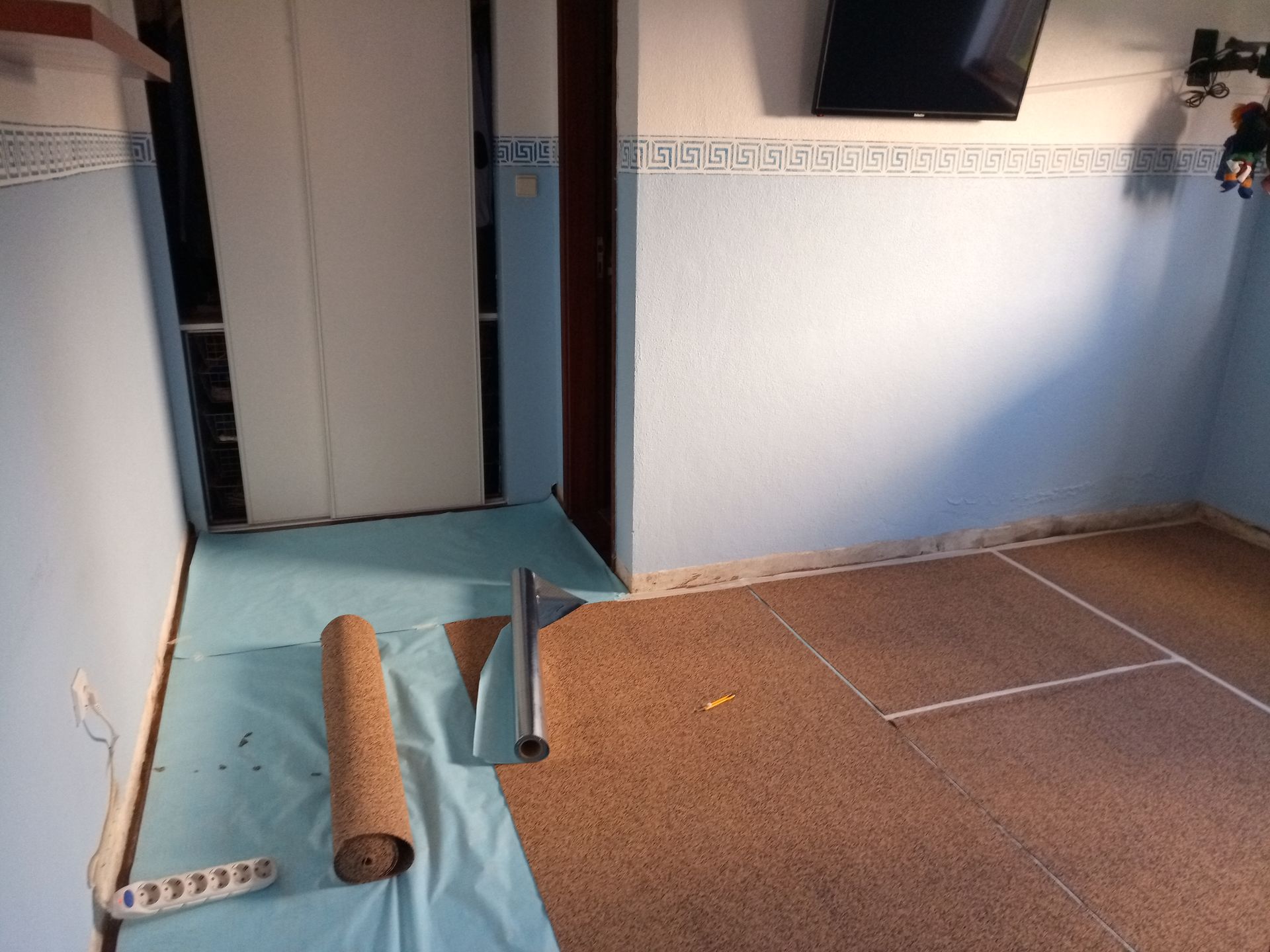 Sala com instalação de piso de cortiça em andamento; paredes azuis, porta, rolo de cortiça e ferramentas visíveis.