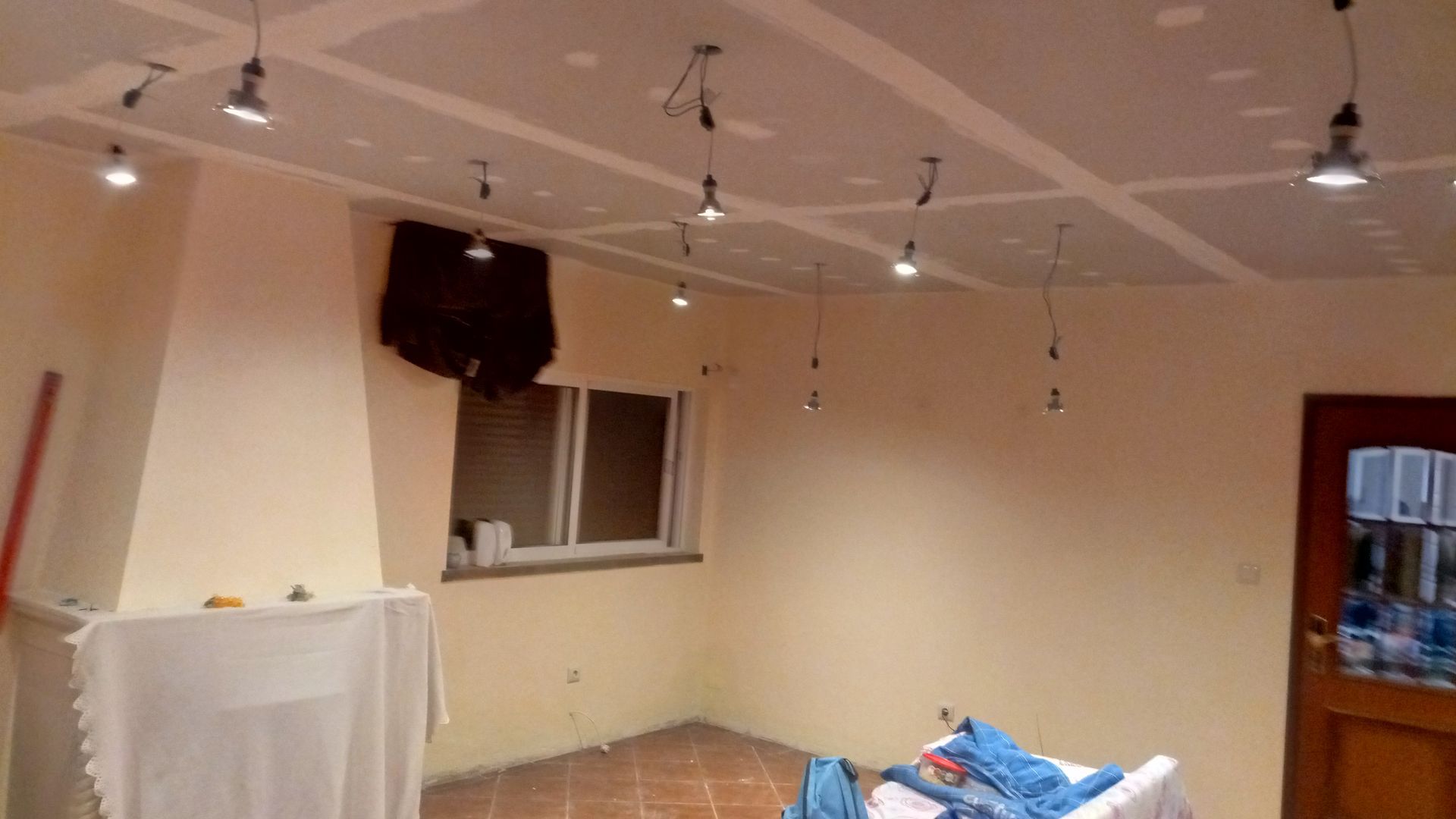 Interior de um cômodo em reforma; teto de gesso acartonado com luminárias pendentes, paredes em tom creme, janela e porta.