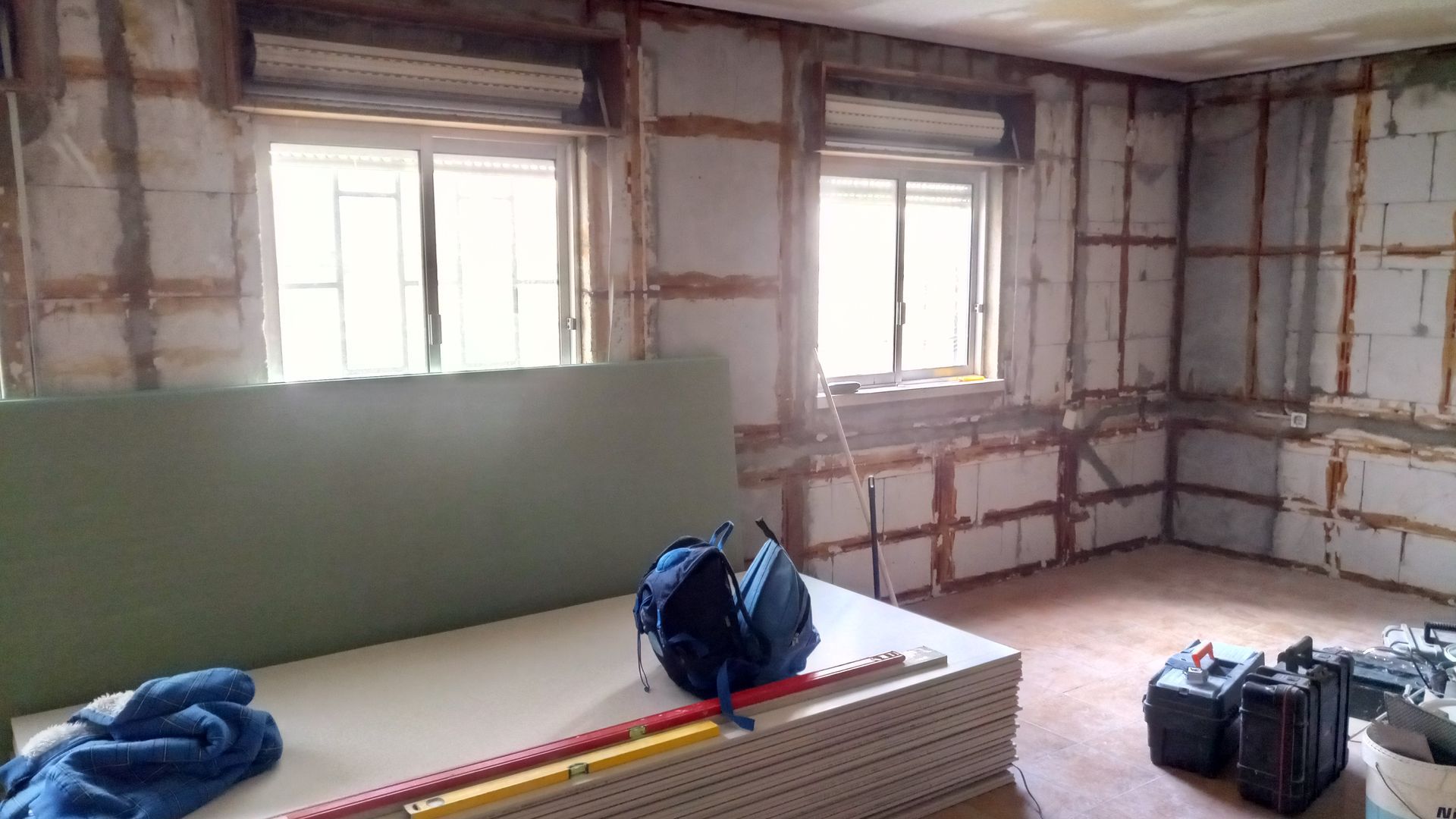 Reforma de cômodo interno com vigas e drywall expostos, duas janelas, ferramentas e uma pilha de drywall.