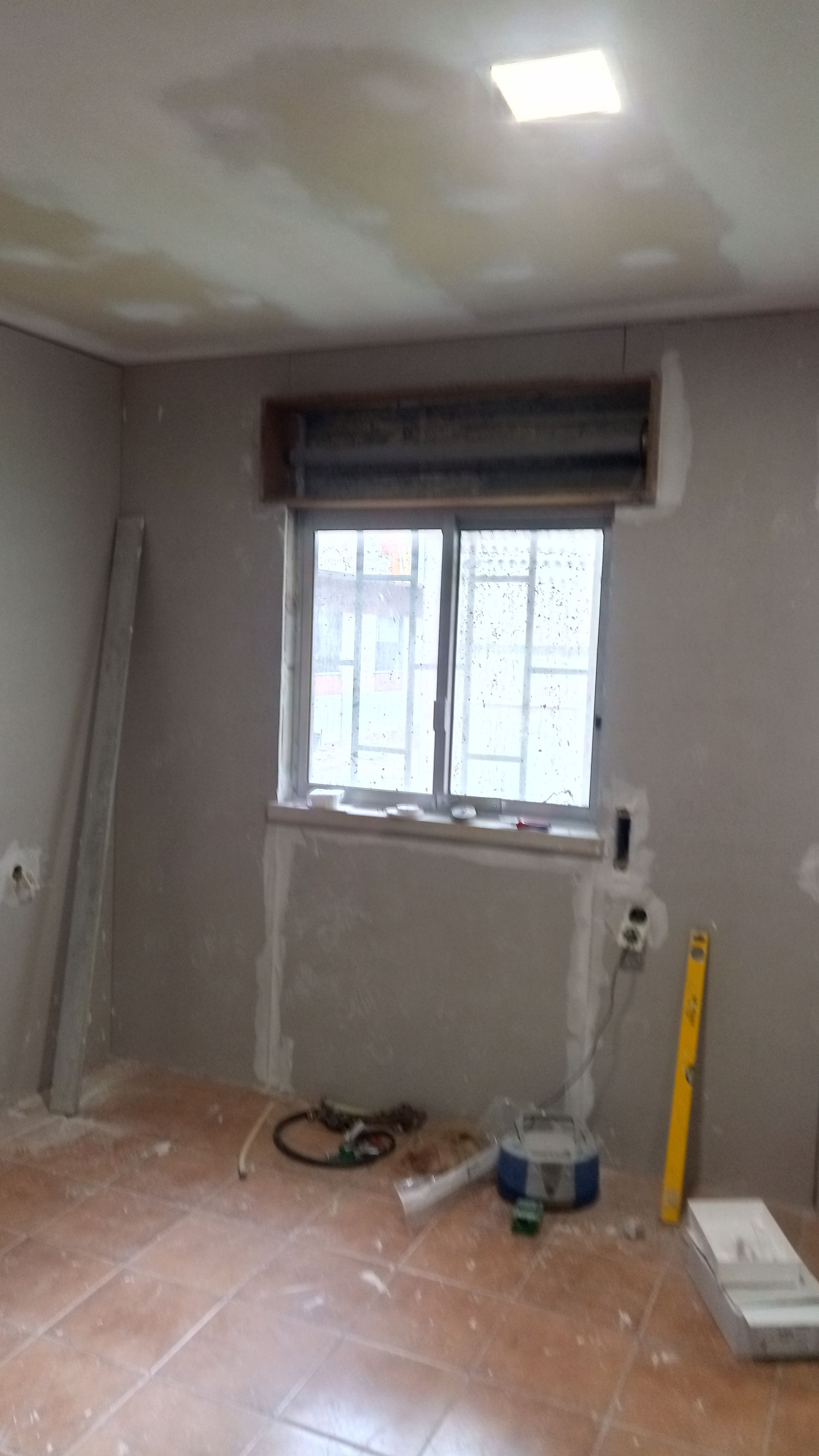 Quarto em reforma com janela, persianas e ferramentas no chão. Paredes e teto inacabados, piso de azulejo.