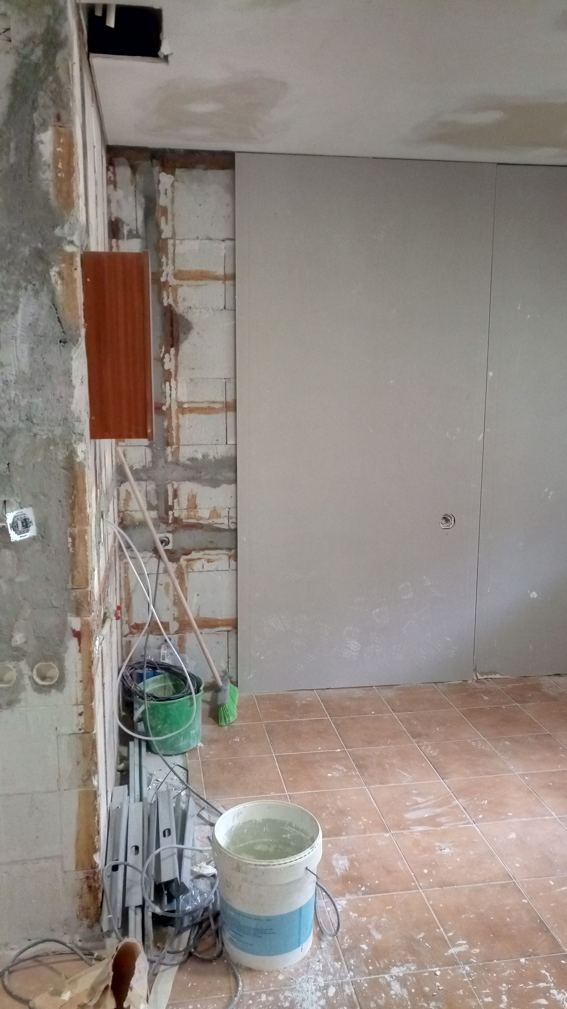 Quarto em reforma com fiação exposta, paredes inacabadas e drywall. Balde e ferramentas no chão.