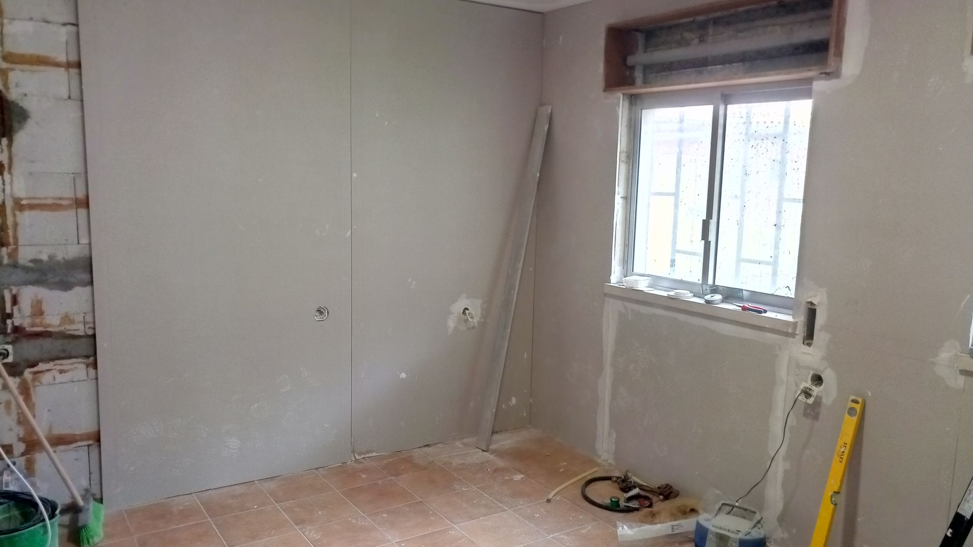 Quarto em reforma: paredes de gesso cartonado, janela, piso sem acabamento, ferramentas presentes.