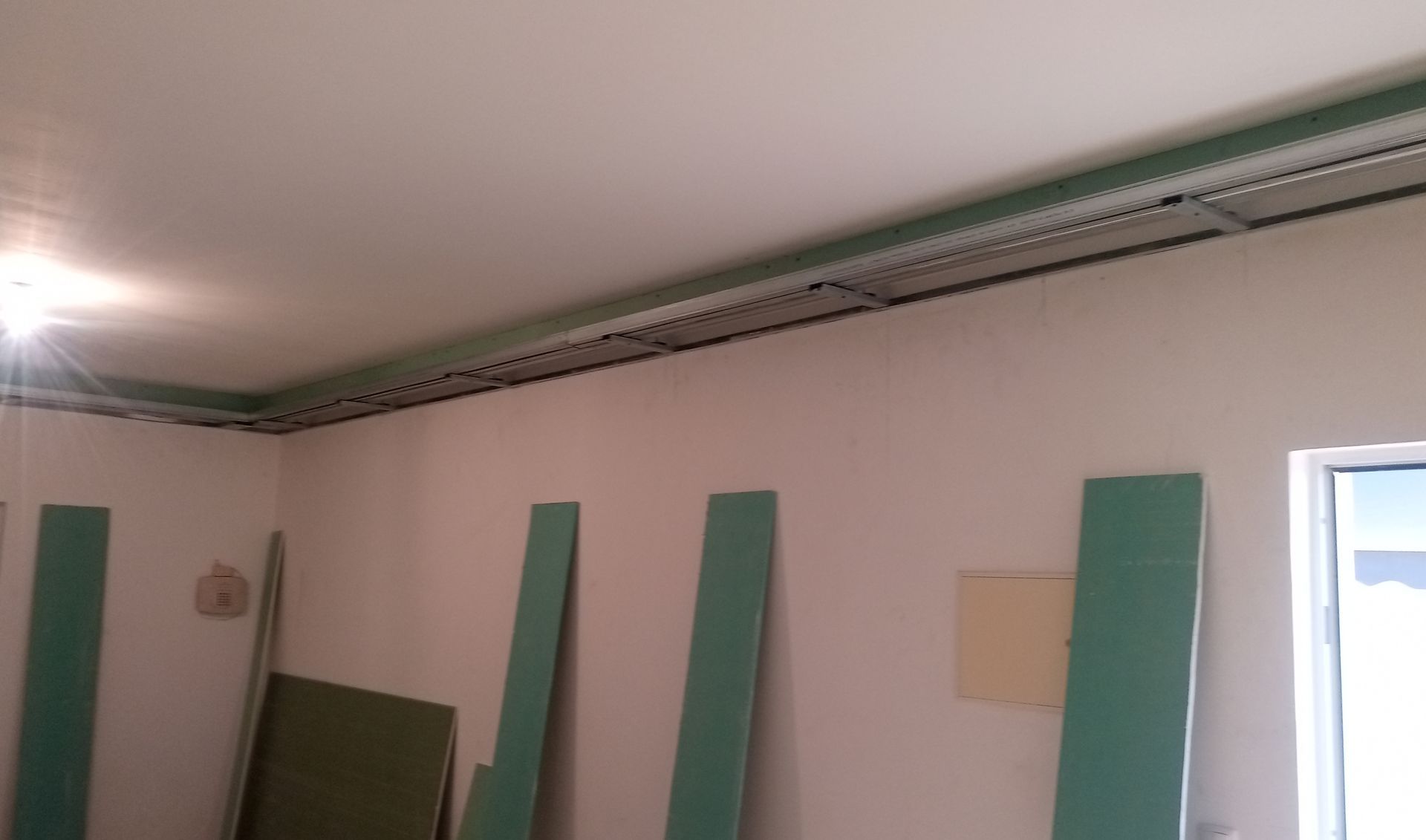 Interior de um cômodo com instalação de drywall inacabada; placas verdes, molduras de teto e janela.