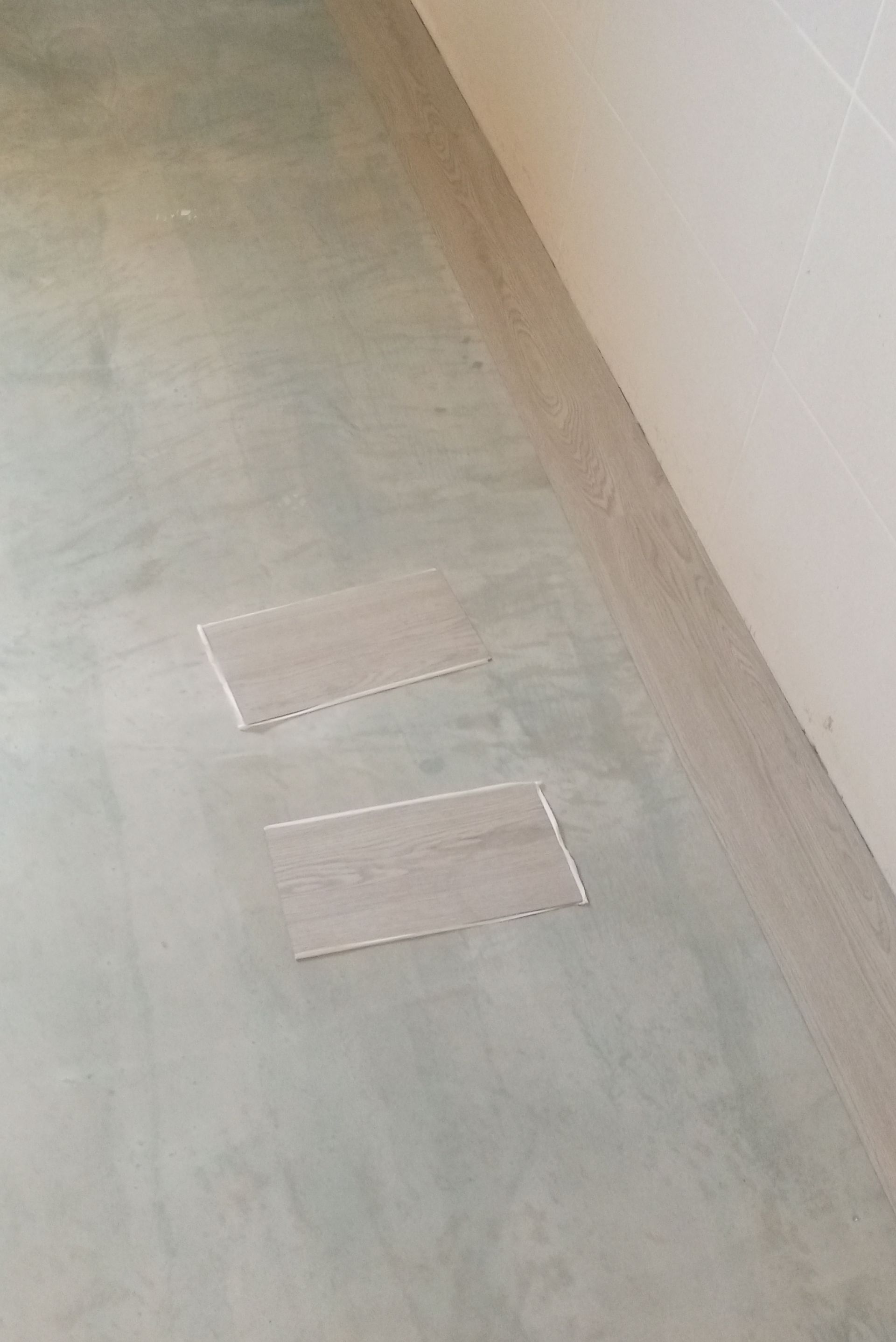 Dois ladrilhos retangulares com aparência de madeira sobre um piso de concreto cinza, próximos a uma parede bege com moldura combinando.
