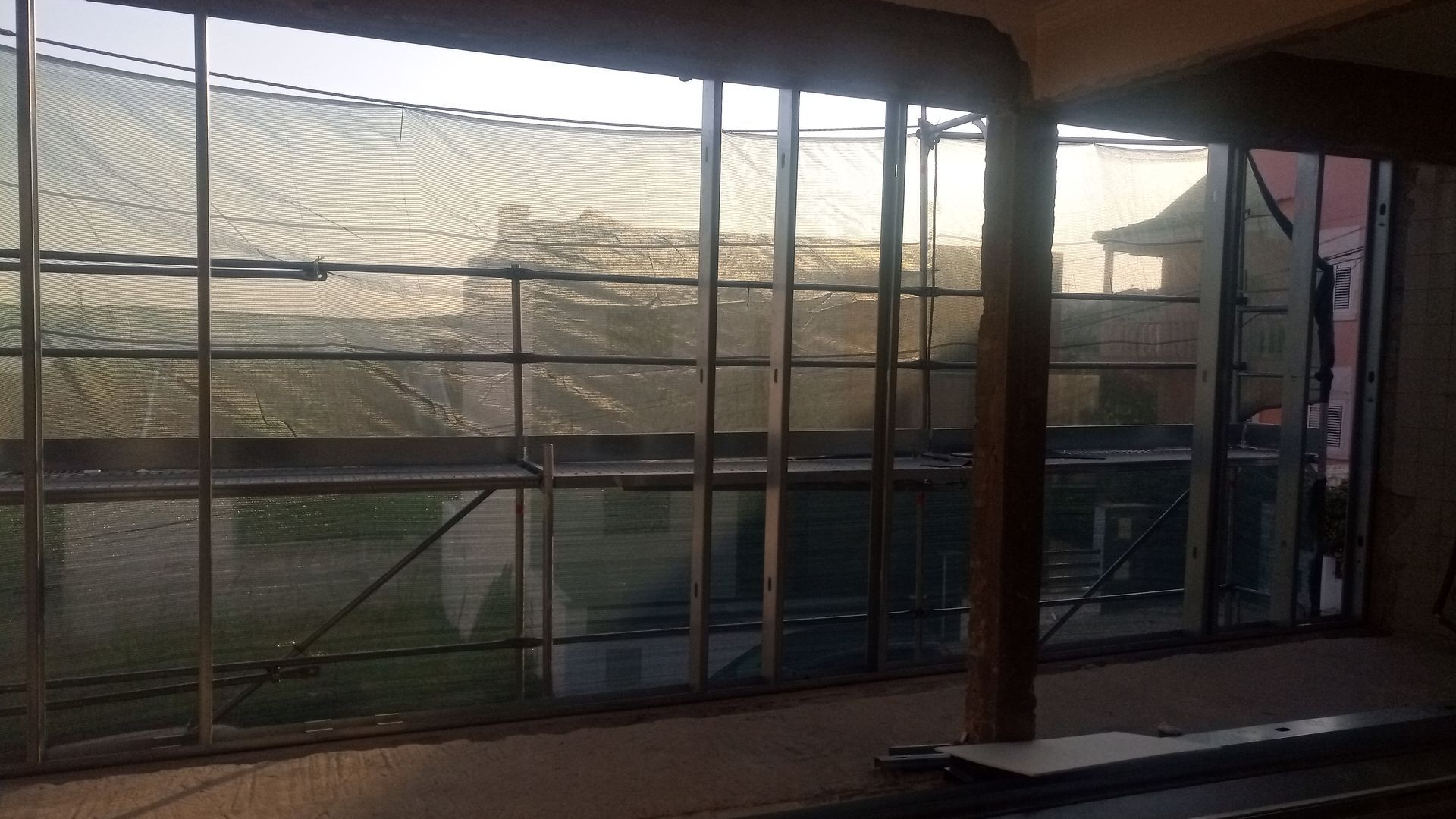 Vista do interior através de uma grande janela, com andaimes do lado de fora, iluminados pelo sol e parcialmente obstruídos por uma cobertura.