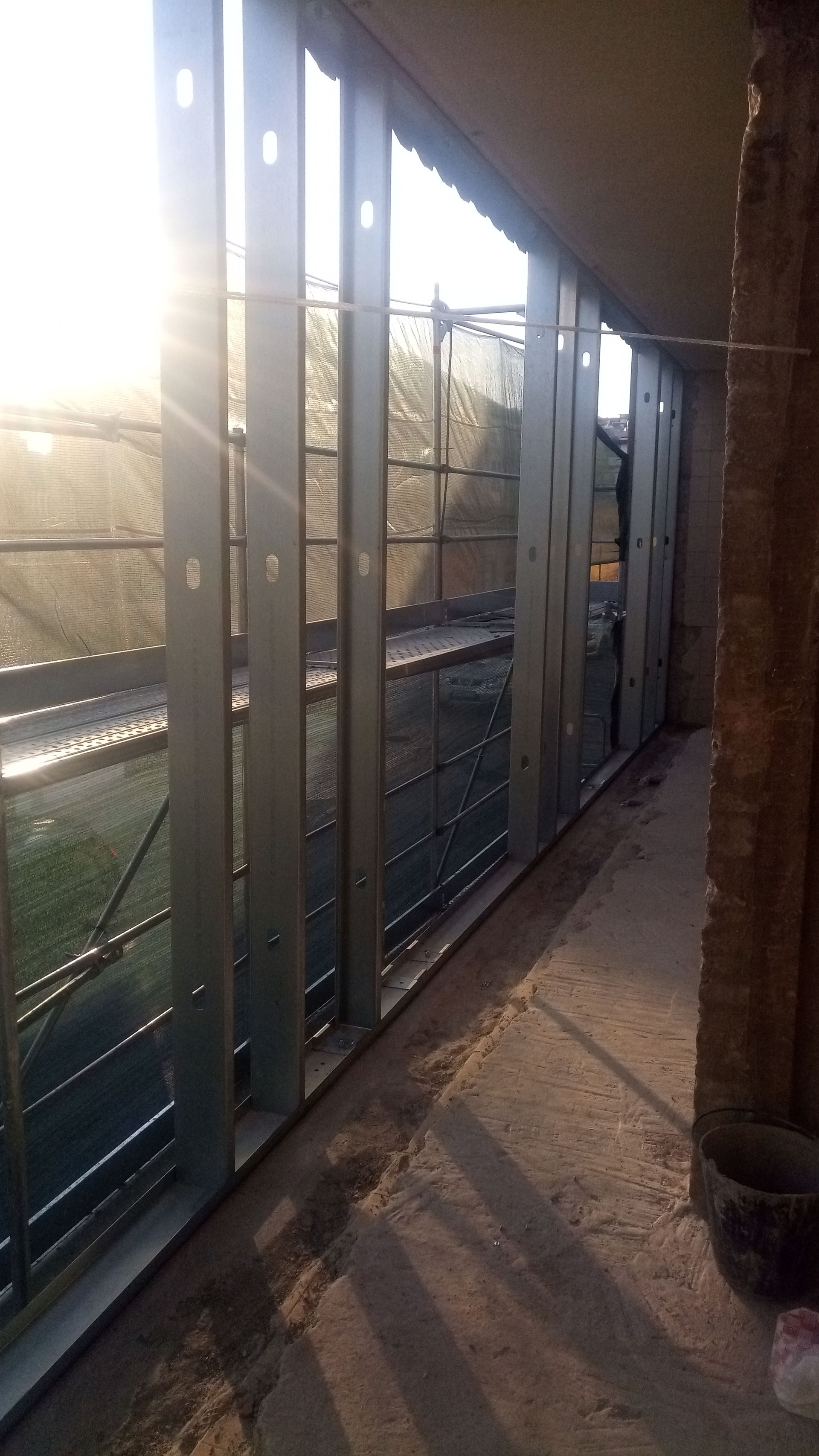 A luz do sol entra por uma janela com moldura de metal e tela mosquiteira. Uma passarela de concreto é visível.