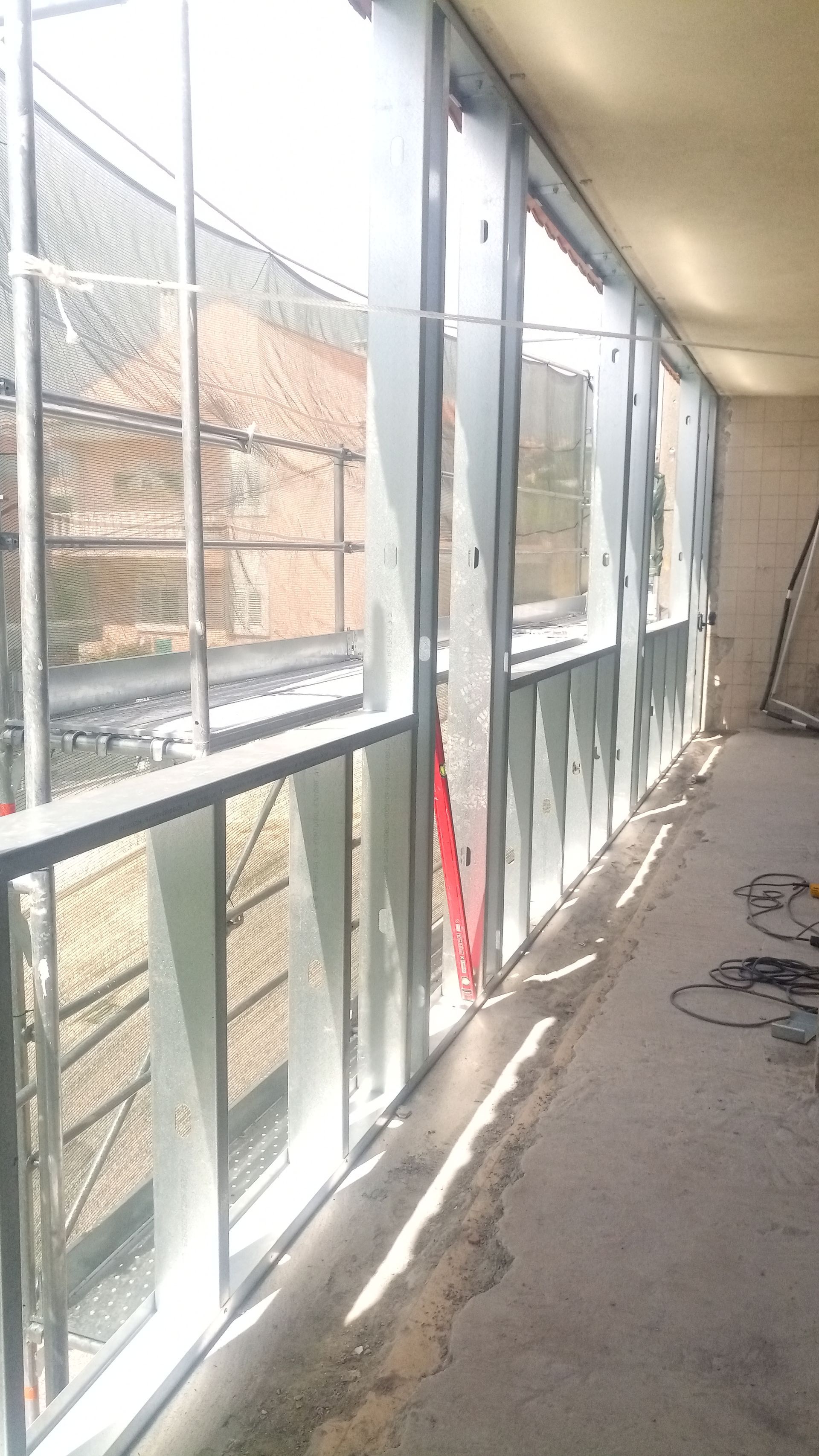 Vista interior de um corredor com janelas de estrutura metálica, construção em andamento, vista para o exterior, luz solar.